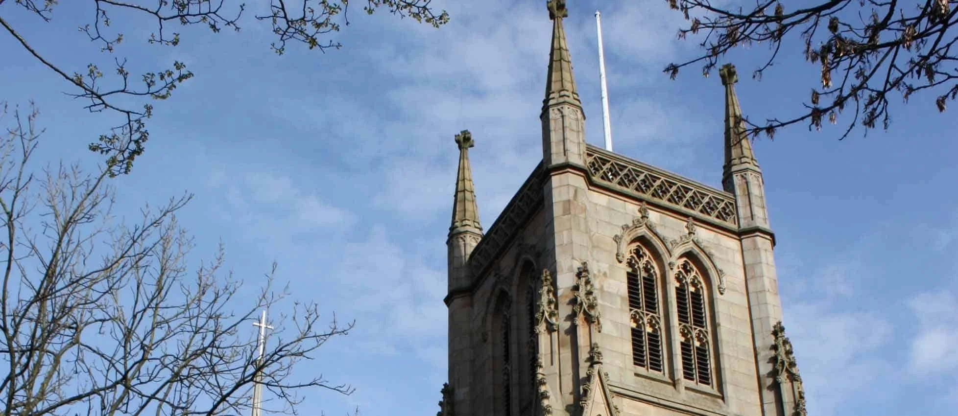 A tall stone church tower with gothic-style windows, decorative spires, and a flagpole on top, set against a background of blue sky with some clouds and tree branches in the foreground.