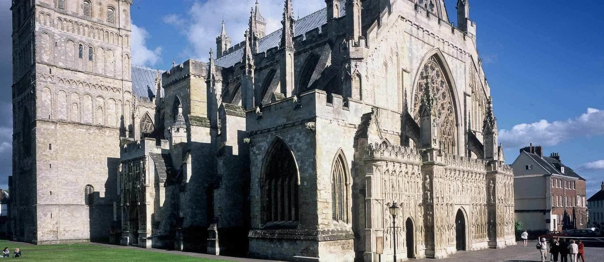 Exterior view of a historic Gothic cathedral with intricate stone carvings, large stained glass windows, and tall spires against a blue sky with some clouds. People are walking and sitting on the grass nearby.
