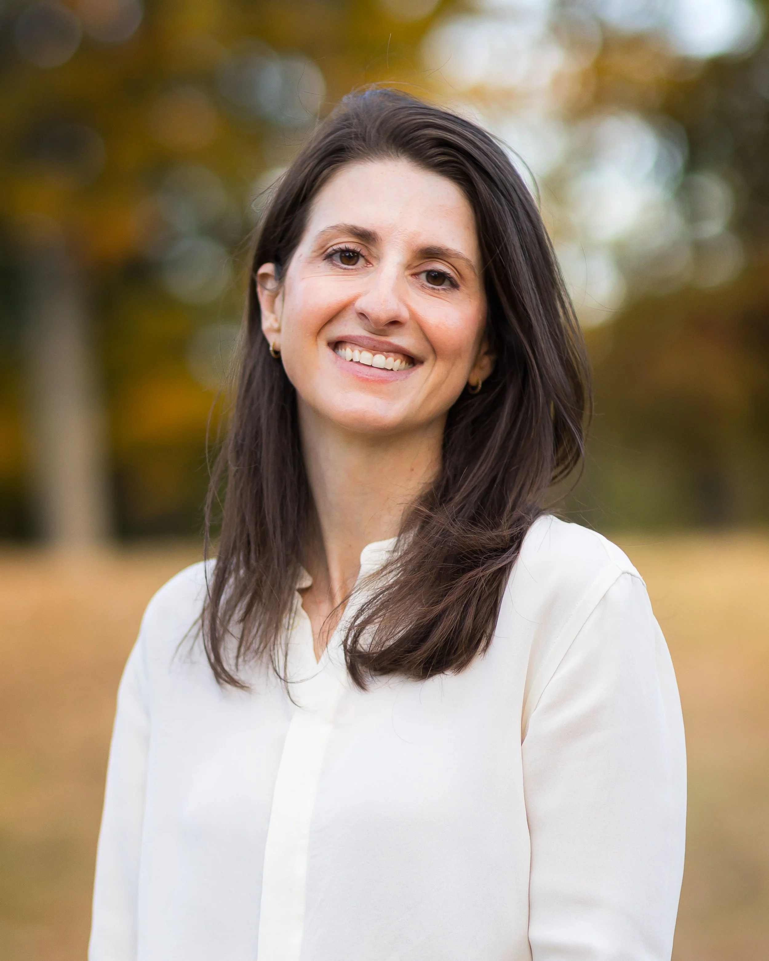 A woman with long dark hair smiling outdoors, wearing a cream-colored blouse, with blurred trees and fall foliage in the background.