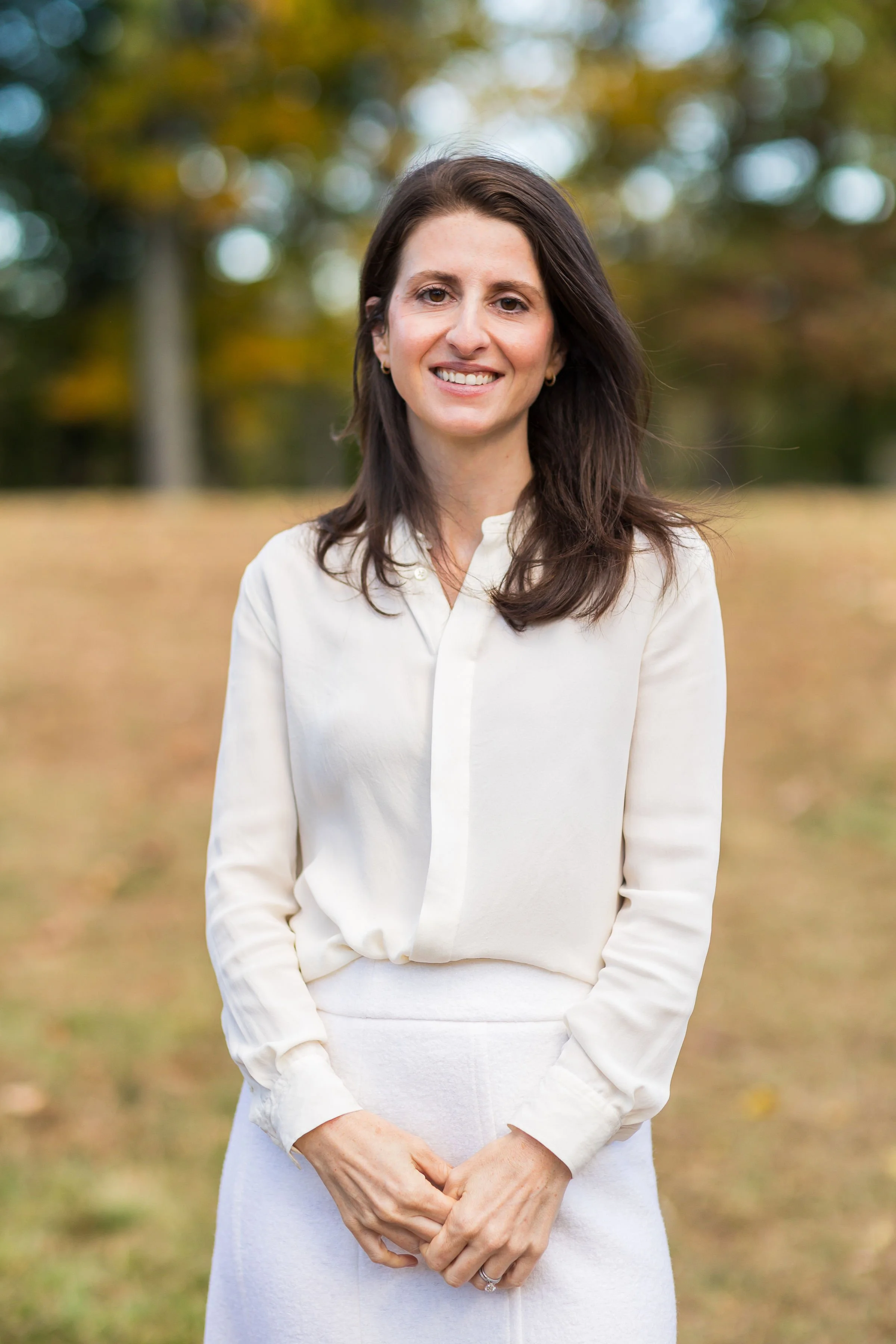 A woman with dark brown hair wearing a white long-sleeve blouse and skirt standing outdoors in a park with autumn foliage in the background.