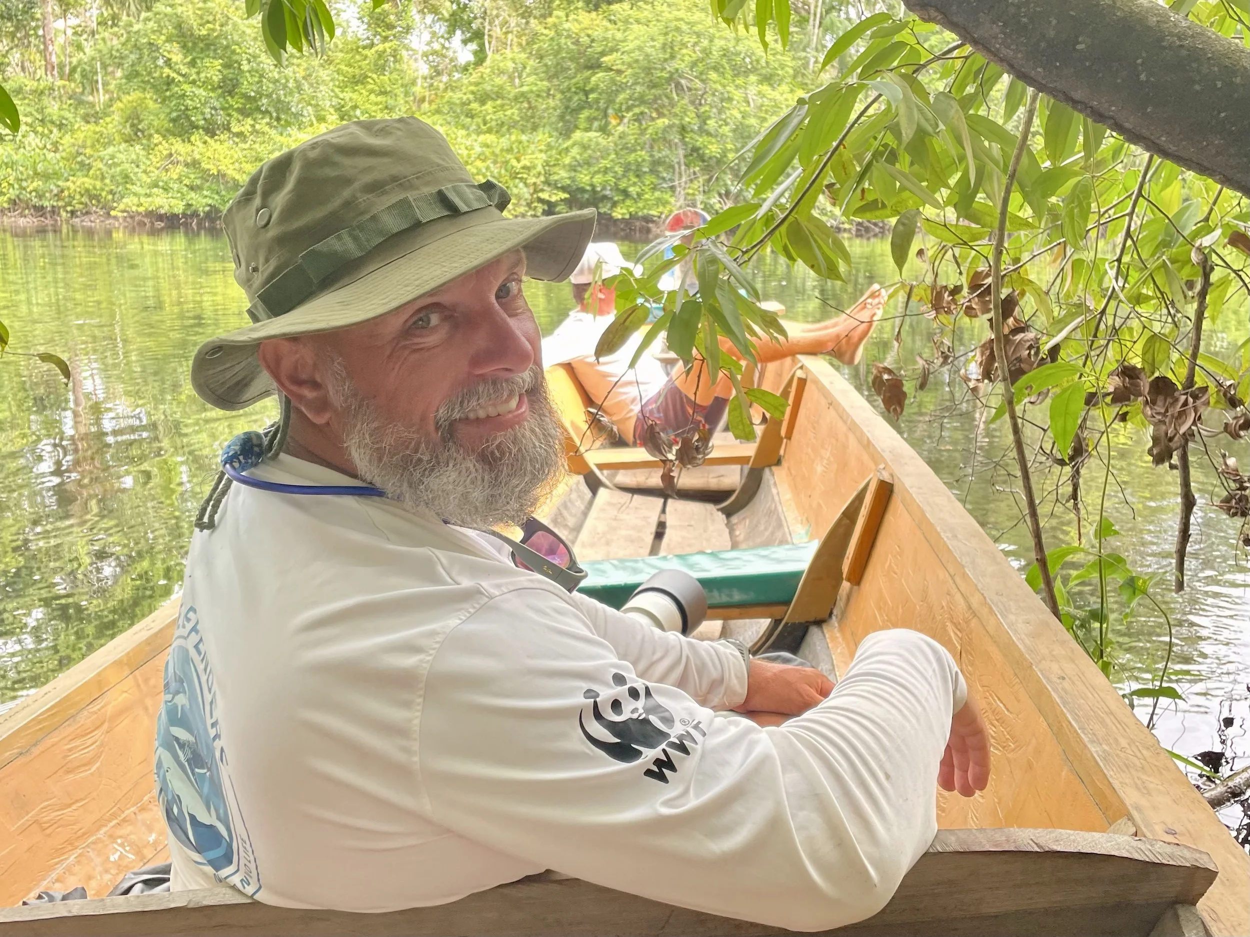 A man with a beard wearing a wide-brimmed hat and a long-sleeve white shirt, smiling while sitting in a wooden boat on a calm river, surrounded by lush green trees and foliage.