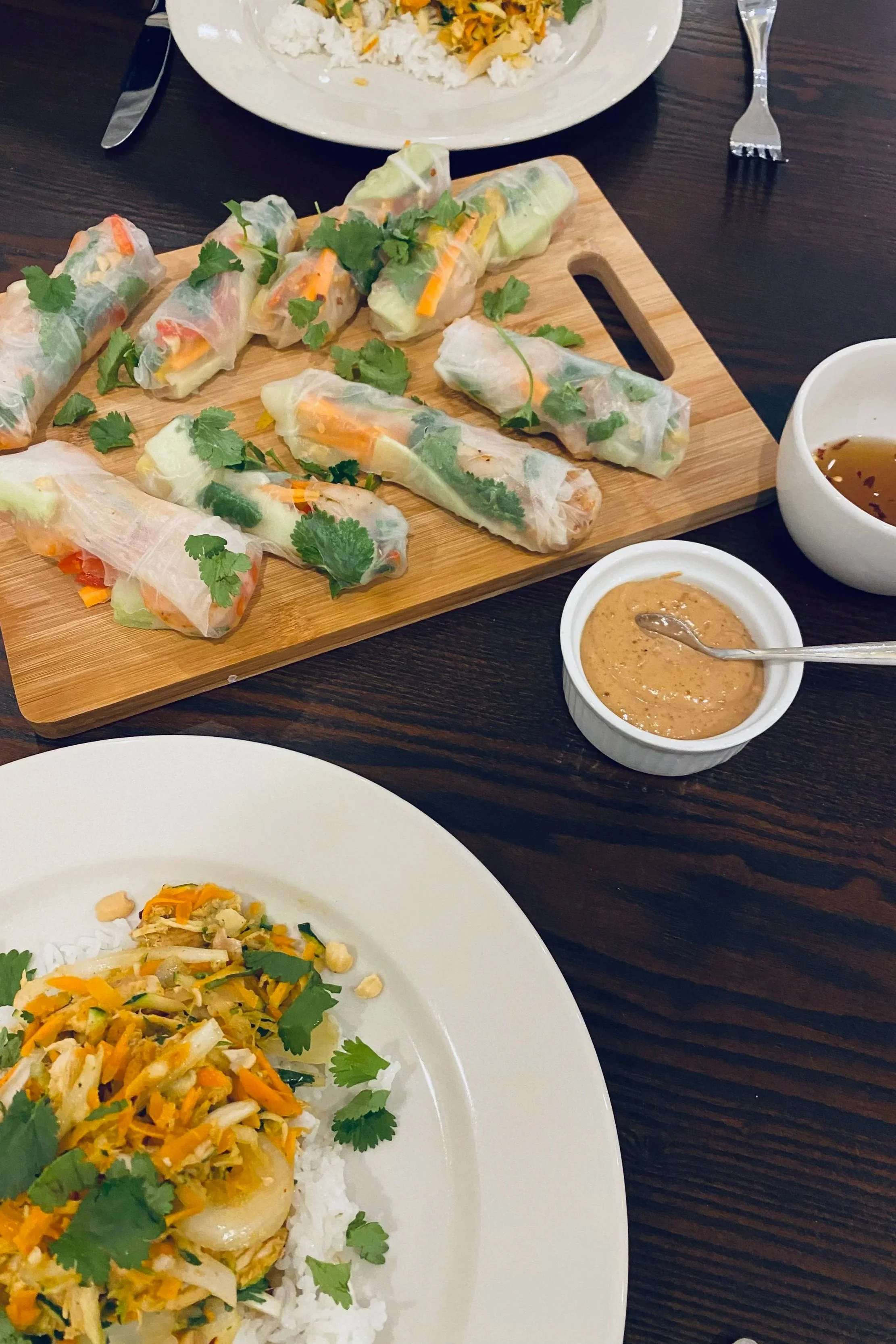 Spring rolls on a wooden cutting board, served with dipping sauces, accompanied by a plate of vegetable stir-fry over rice, on a dark wood table.