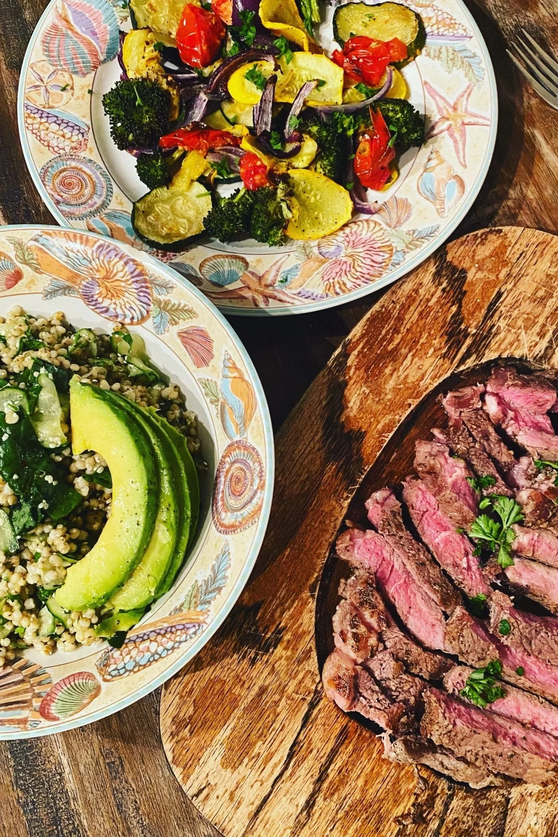 A plate of mixed roasted vegetables including zucchini, cherry tomatoes, red onions, and broccoli. A bowl of tabbouleh salad with cucumber slices and avocado. A wooden board with sliced grilled steak garnished with herbs.