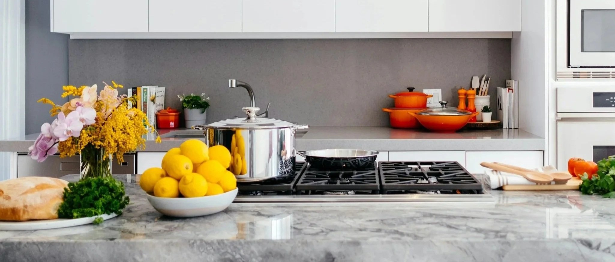 Modern kitchen with marble countertop, bouquet of flowers, lemons in a bowl, bread, parsley, orange pots, cutting boards, and various cooking utensils.