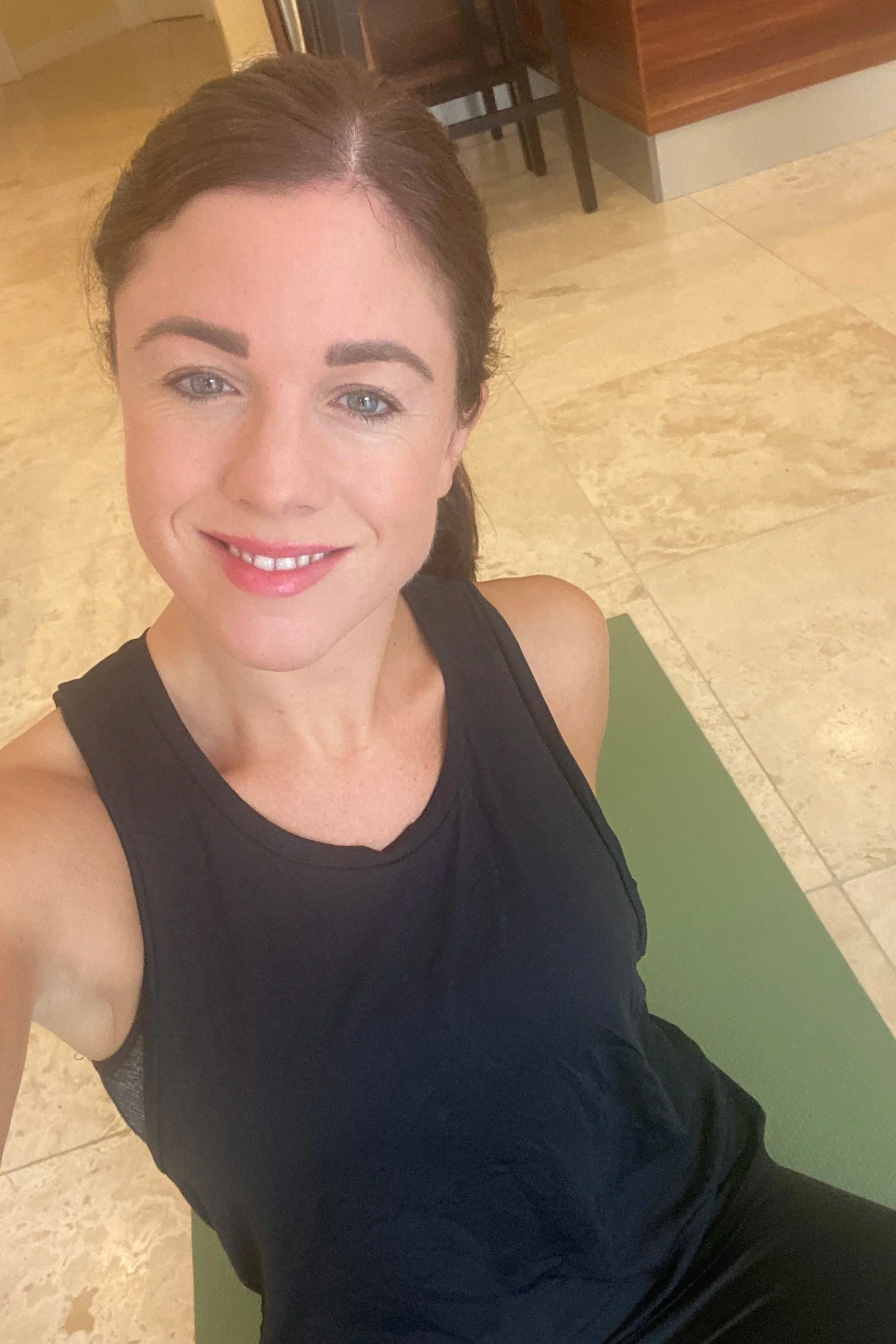 A woman with brown hair pulled back, wearing a black sleeveless athletic top, smiling and sitting on a green yoga mat in an indoor gym or fitness center.