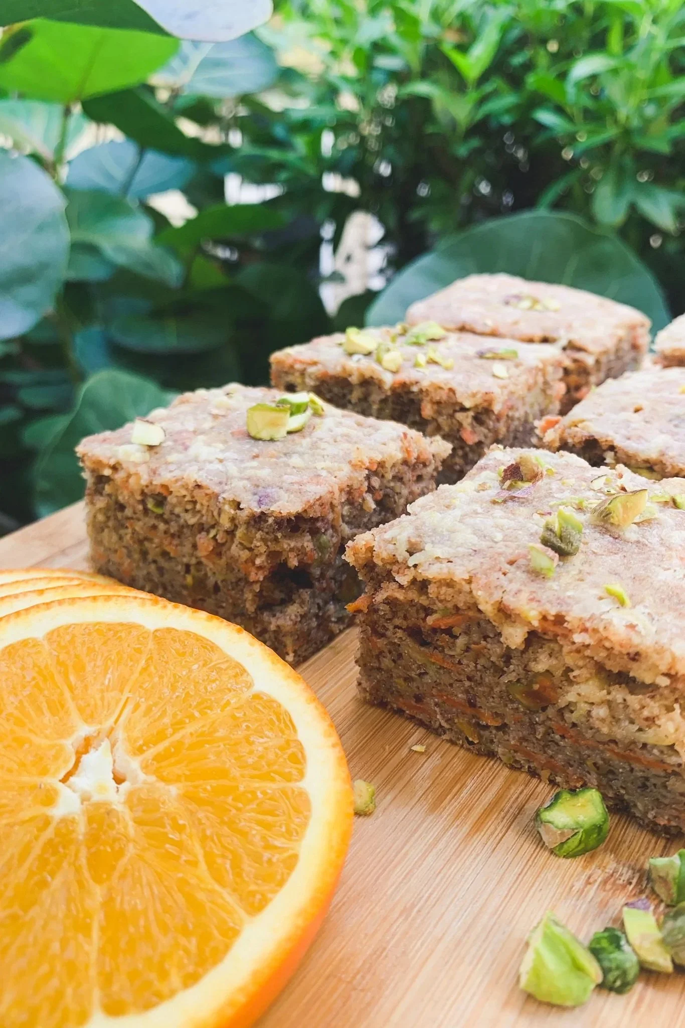 Close-up of sliced orange and square pieces of pistachio cake on a wooden surface with green foliage in the background.