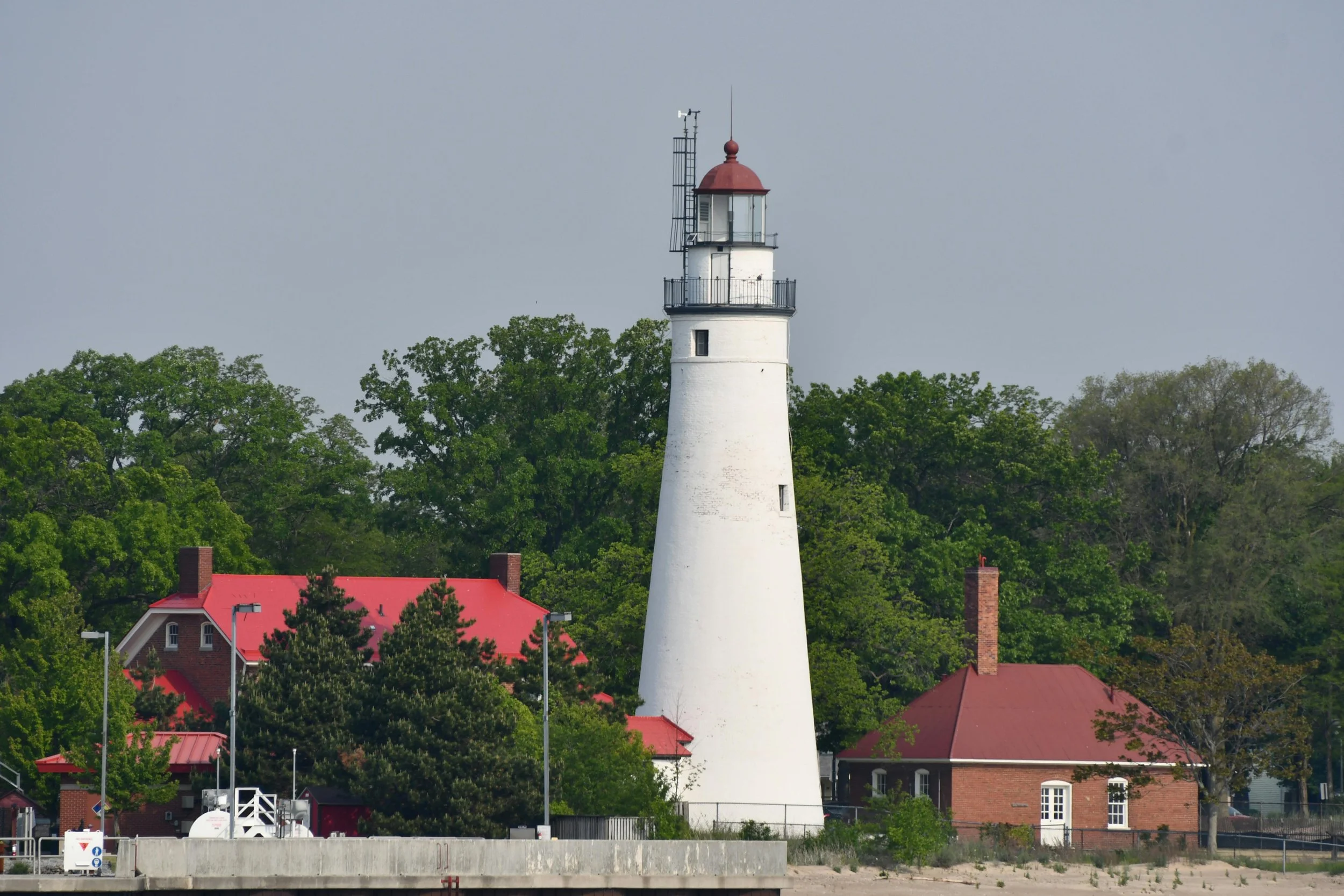 A white lighthouse with a red top, surrounded by green trees and red-roofed buildings.