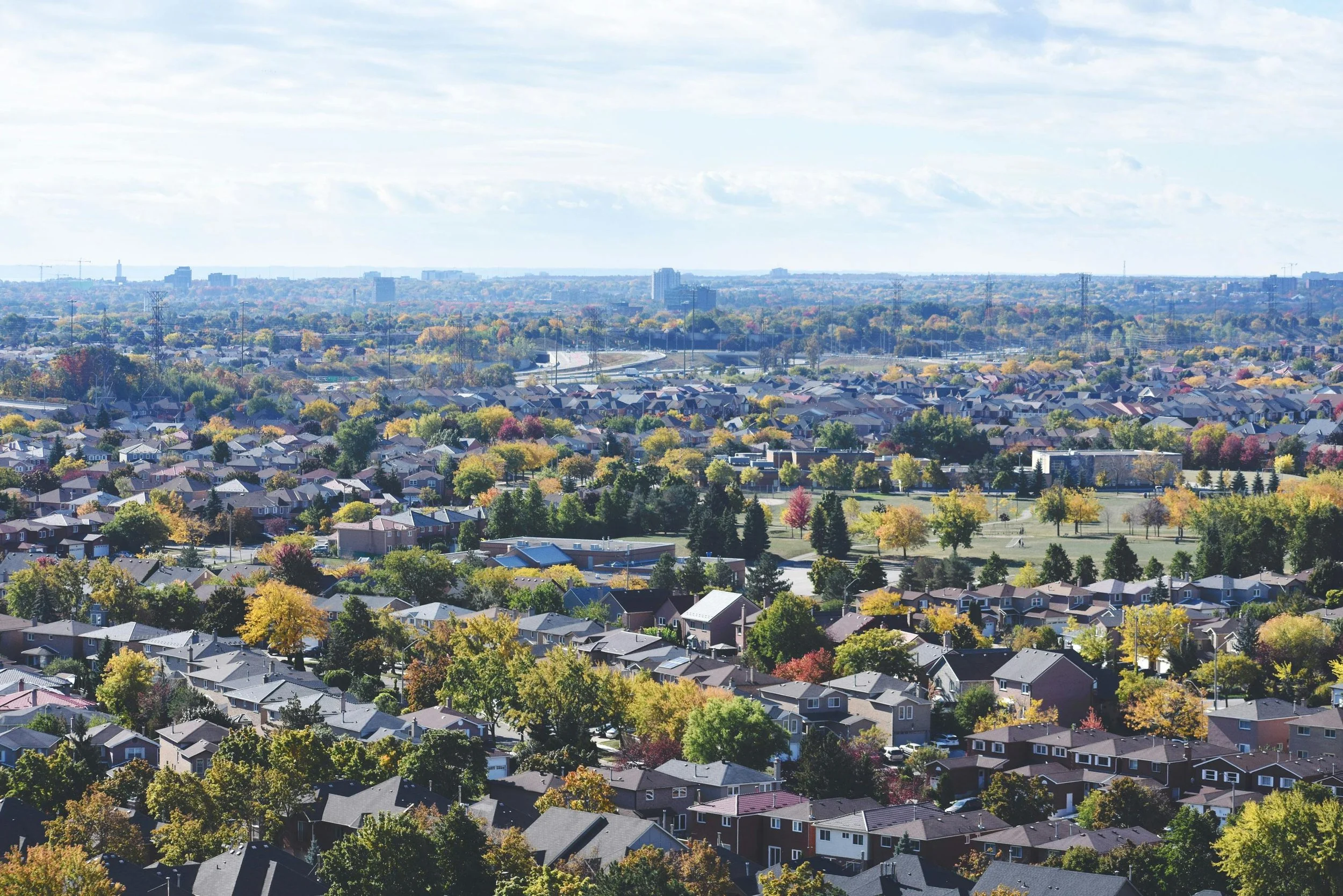Aerial view of a suburban neighborhood with numerous houses, trees with fall foliage, roads, and distant city skyline under a partly cloudy sky.
