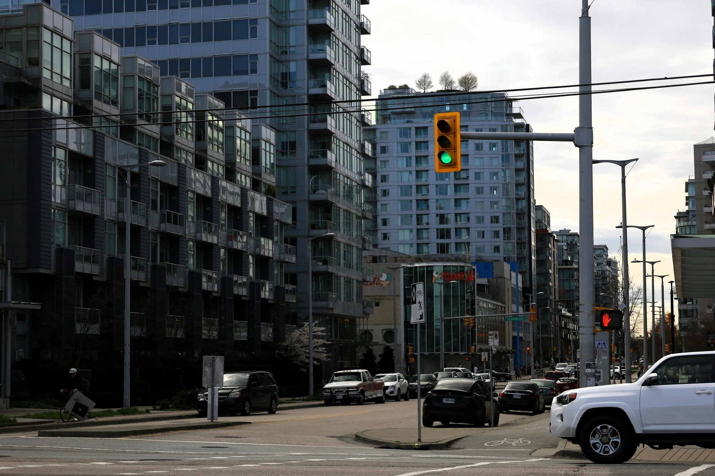 A city street scene with tall modern glass apartment buildings, a traffic light showing green, parked cars along the street, and a bicycle lane with a bike symbol. Overcast sky.