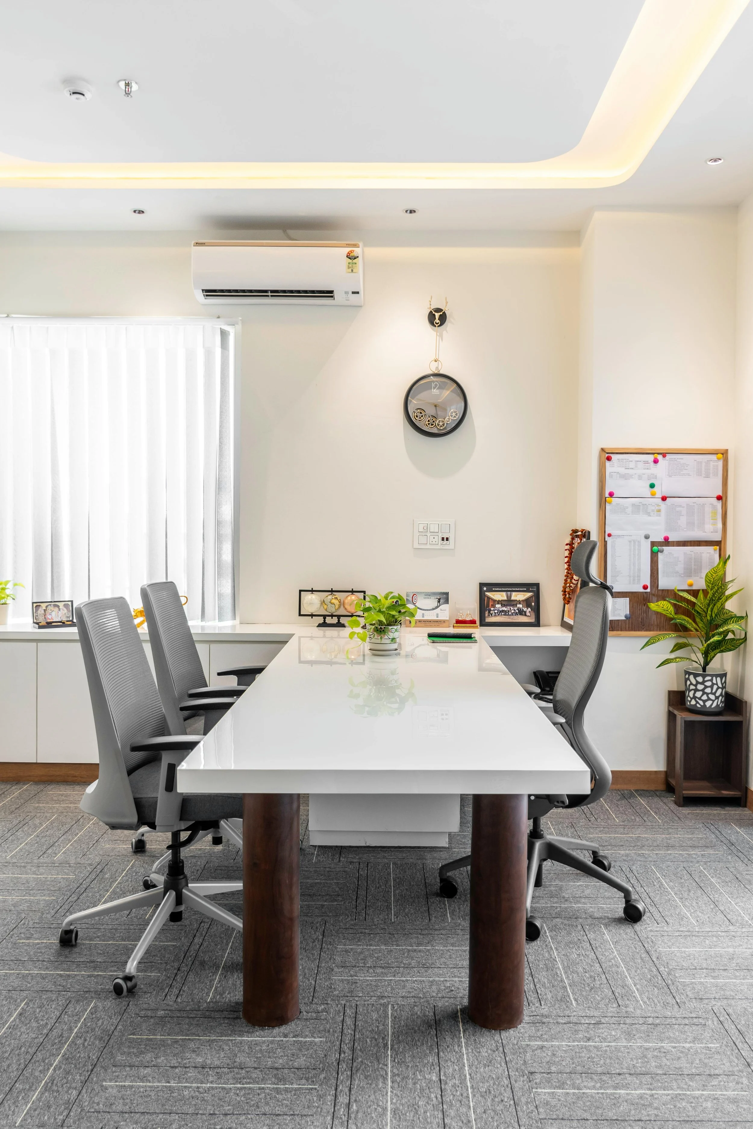 An office conference room with a white table, gray office chairs, a wall clock, potted plants, framed photographs, and a bulletin board with papers, overhead lighting, a window with vertical blinds, and an air conditioning unit.