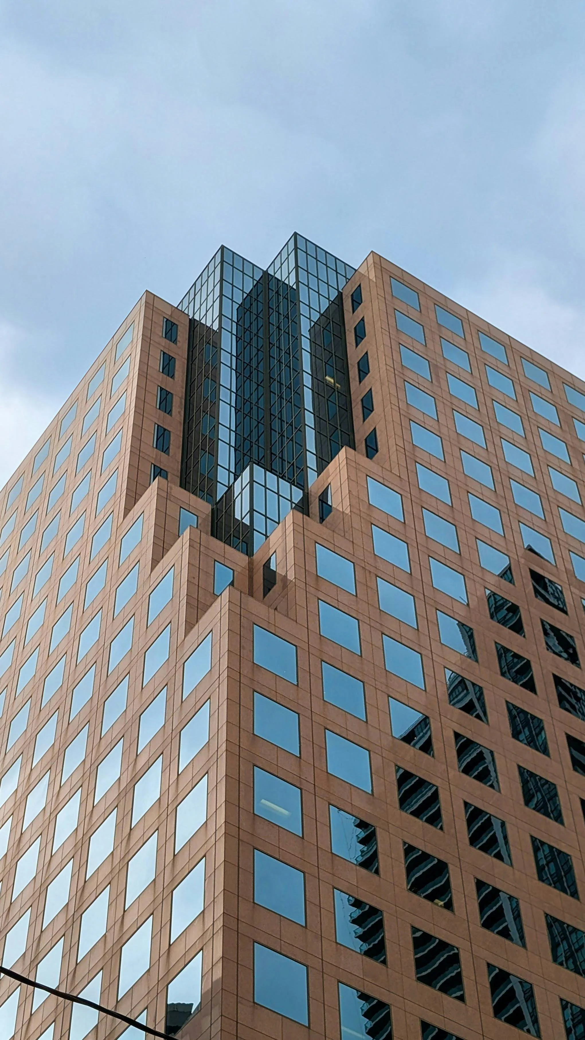 A tall modern building with a facade of brownish-orange panels and numerous blue-tinted windows, topped with a section with reflective glass windows, against a cloudy sky.