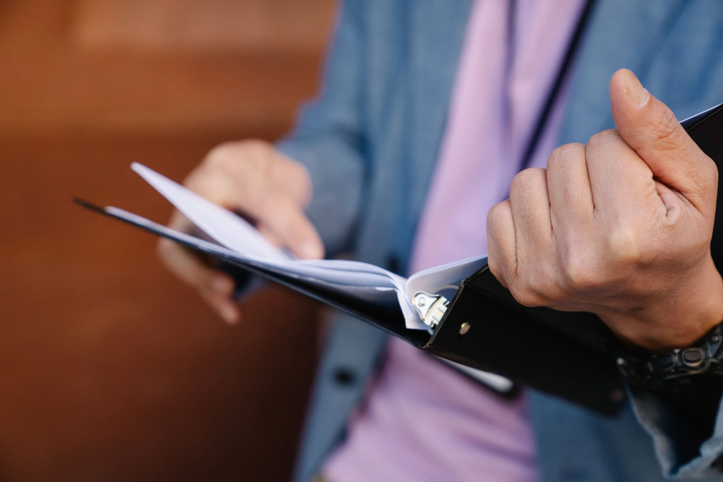 Close-up of a person holding a smartphone in one hand and a notebook or planner in the other, with a blurred brown background.
