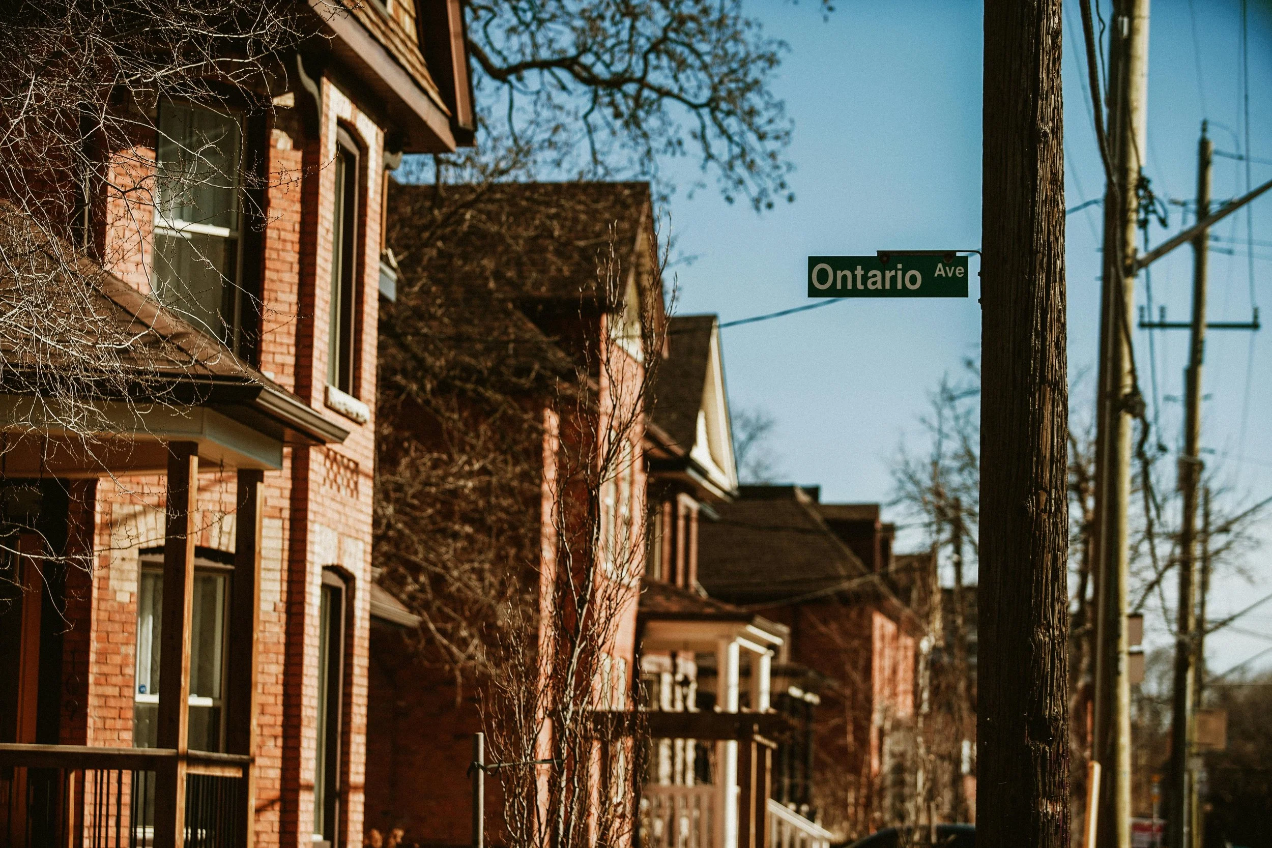 Street sign reading 'Ontario Ave' attached to a wooden pole on a residential street with brick houses and leafless trees.