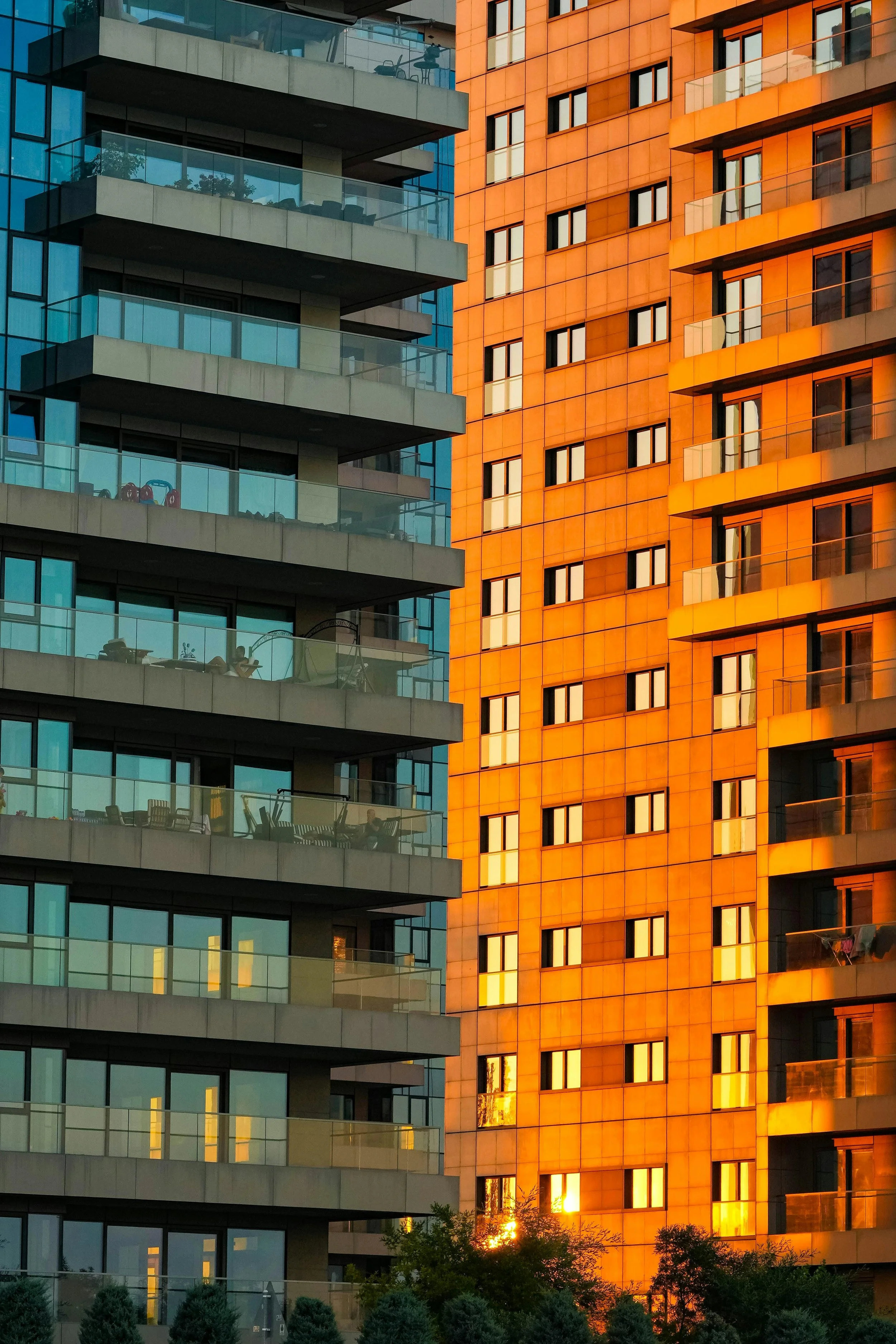 A cityscape featuring two modern high-rise buildings. The building on the left has a blue glass exterior with glass balconies, while the building on the right has an orange tinted exterior with many windows. The sunlight is reflecting off the buildings, creating a warm glow at the bottom of the image.