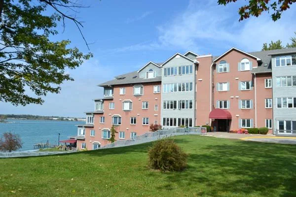 Multi-story apartment building with pink and gray exterior, overlooking a body of water, with a grassy lawn and trees in the foreground.