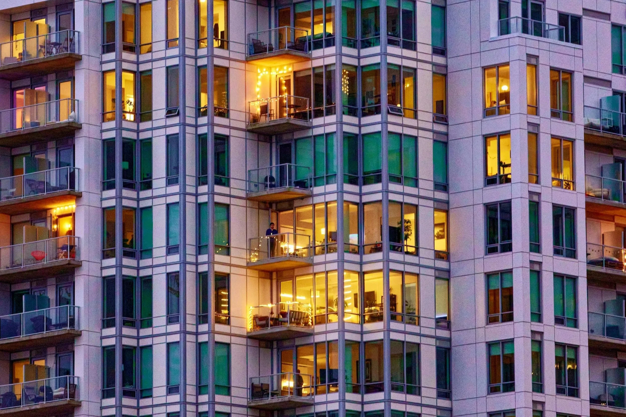 A tall apartment building with lit windows and small balconies, some with visible furniture and decorations.
