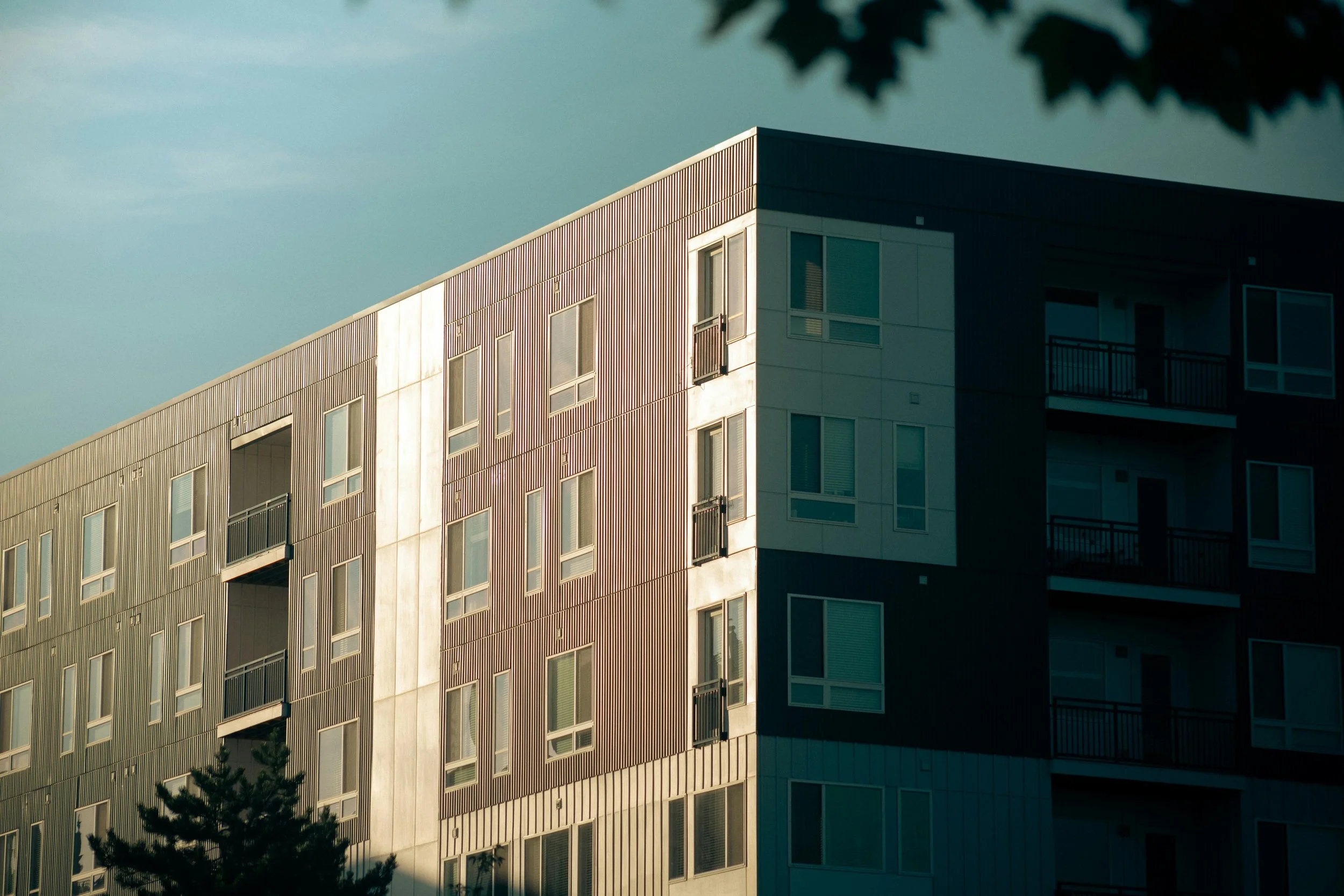 Multi-story modern apartment building with balconies and windows, partly in shadow, with tree leaves at the top of the image.