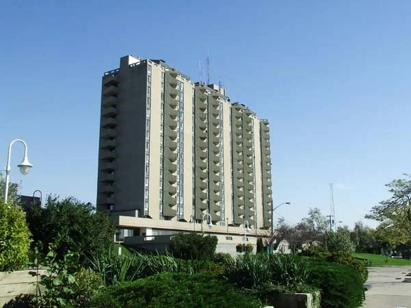 Tall multi-story apartment building with balconies, surrounded by greenery and street lamps under a clear blue sky.