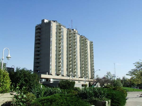 A tall, modern apartment building with multiple balconies, situated in a park-like area with greenery, trees, and a clear blue sky.