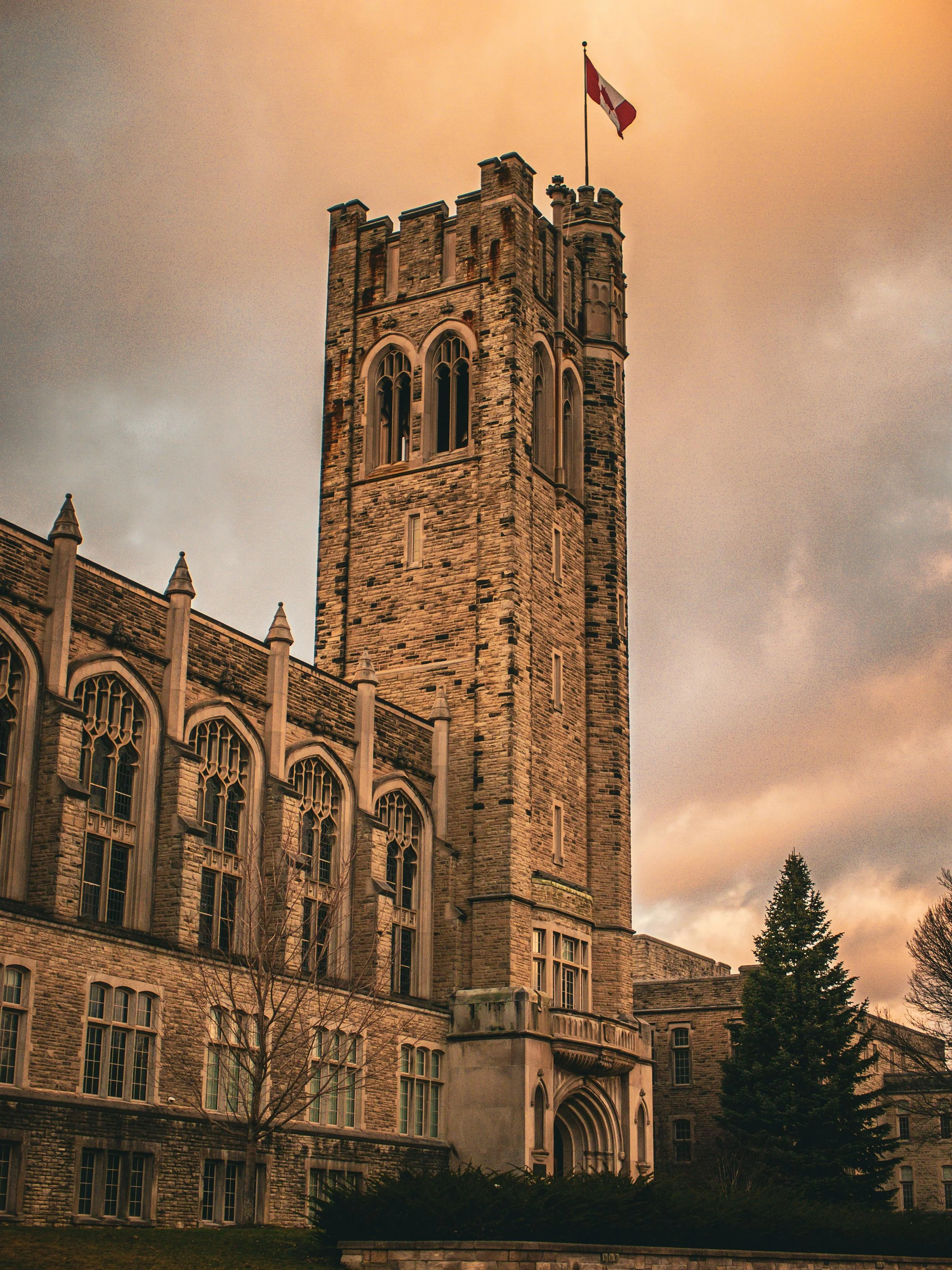 A tall stone castle-like building with a turret and a Canadian flag on top, set against a cloudy sky at sunset.