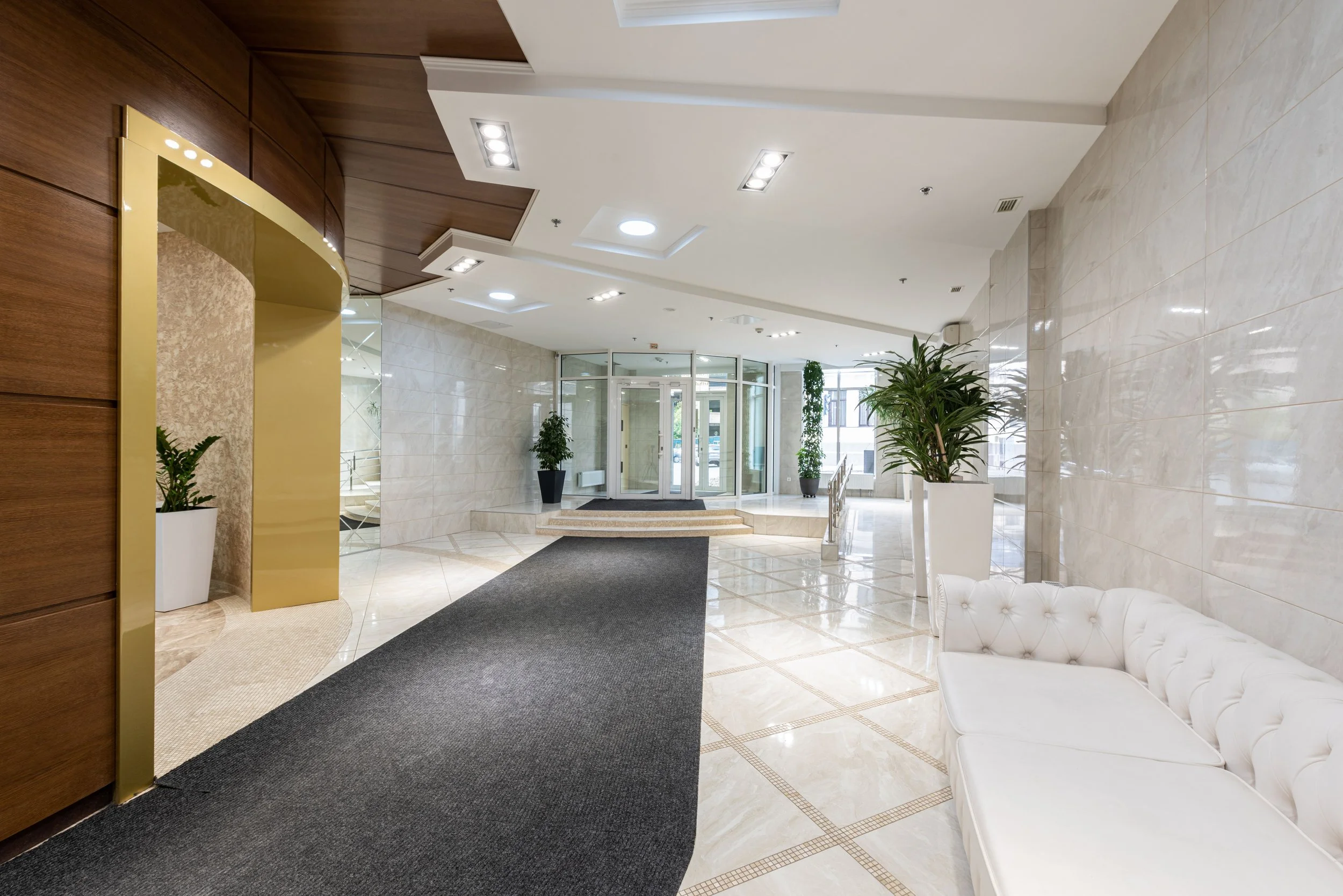 Lobby area with white marble walls and floor, potted plants, white tufted bench, and a revolving glass door entrance.