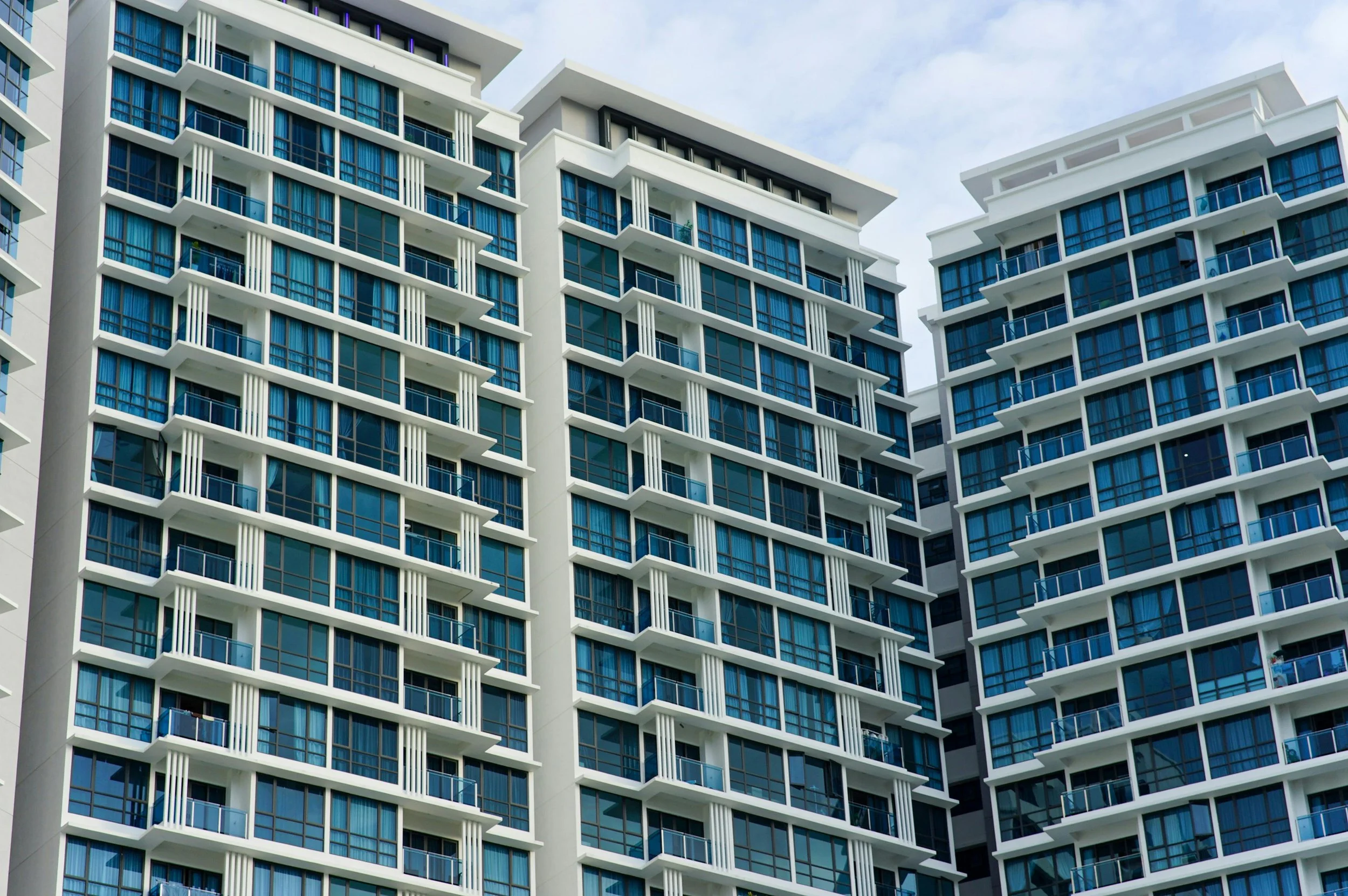A close-up view of a modern high-rise apartment building with multiple floors, featuring glass windows, blue curtains, and small balconies.