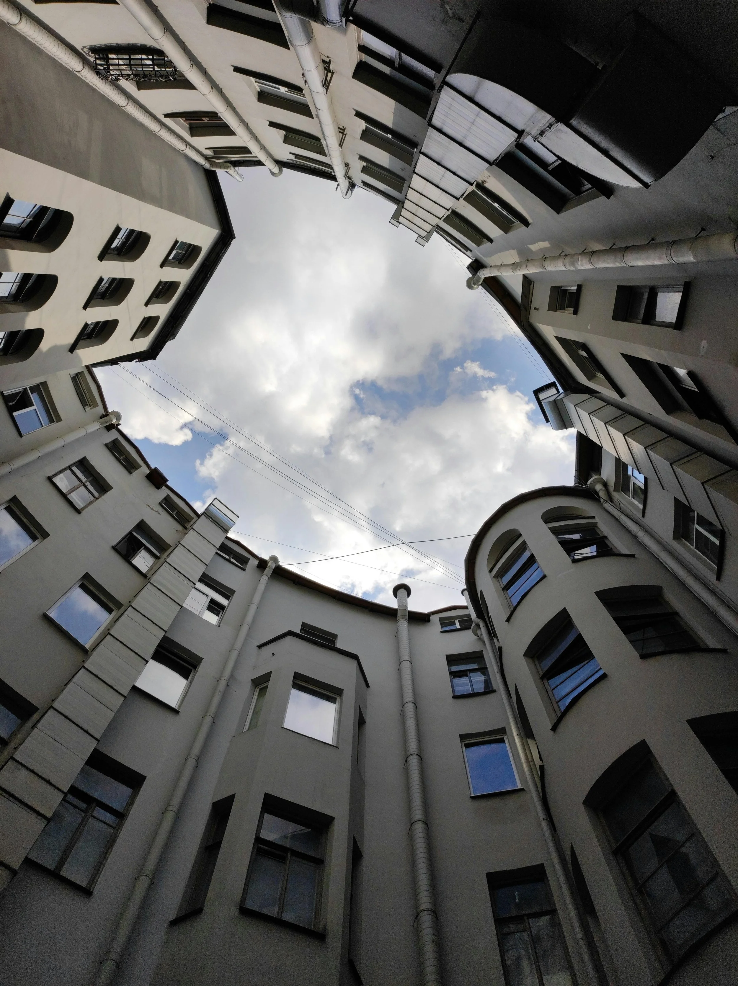 Looking up into a circular courtyard of a multi-story building with white walls, multiple windows, and pipes on the exterior, with a partly cloudy sky visible overhead.