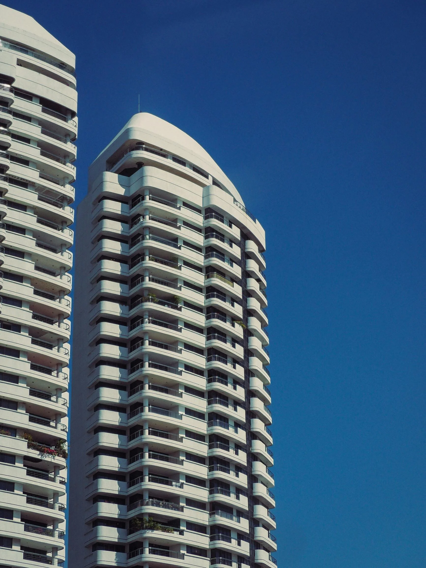 Two modern white high-rise buildings with multiple balconies against a clear blue sky.