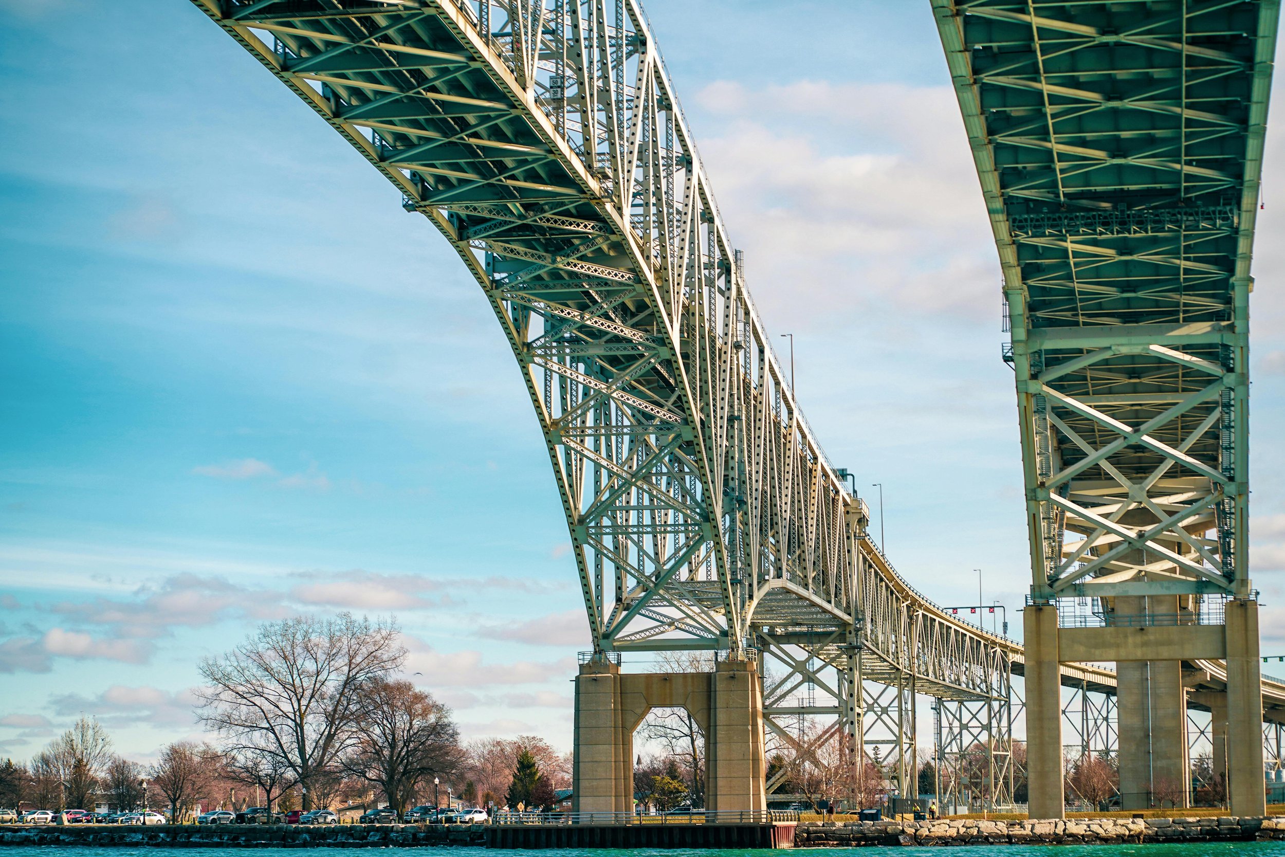 View of a large green steel bridge spanning over water, with parking lot and leafless trees in the background, under a partly cloudy sky.