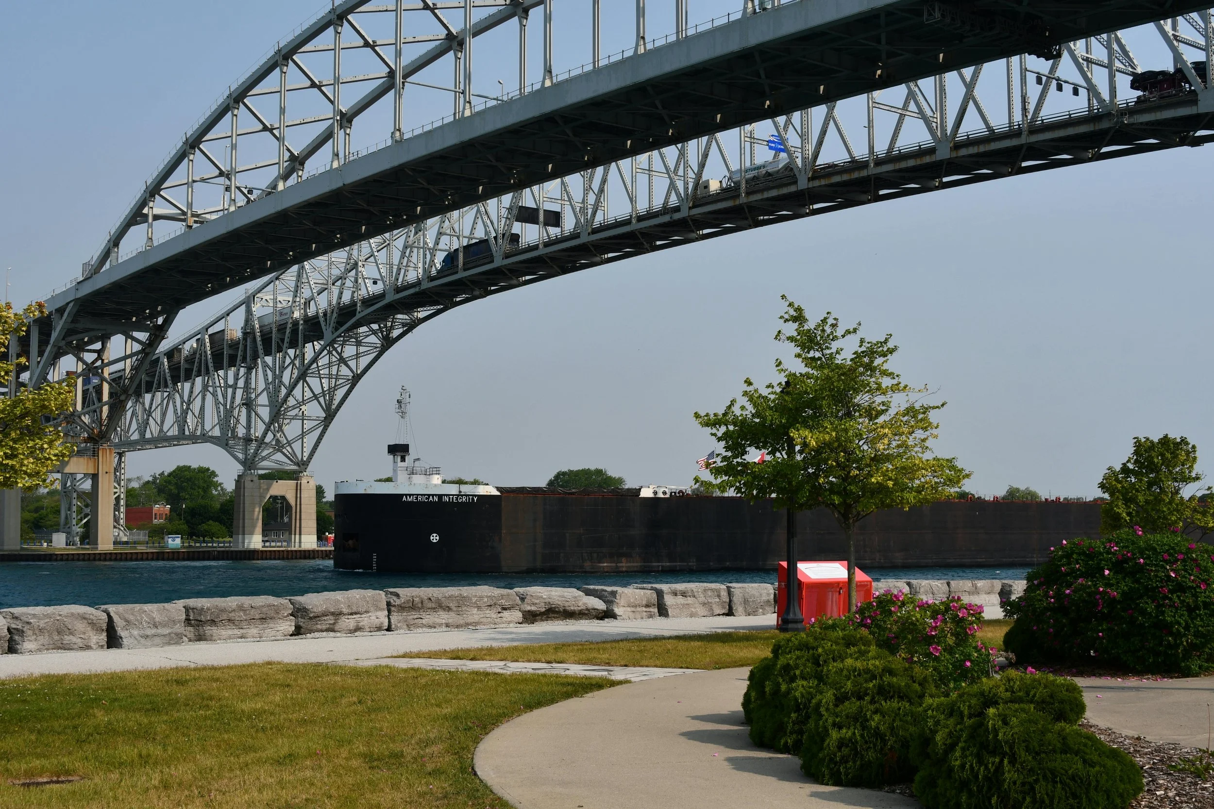 View of a large black cargo ship named 'American Integrity' passing under a steel arch bridge over water, with a park and flowering bushes in the foreground.