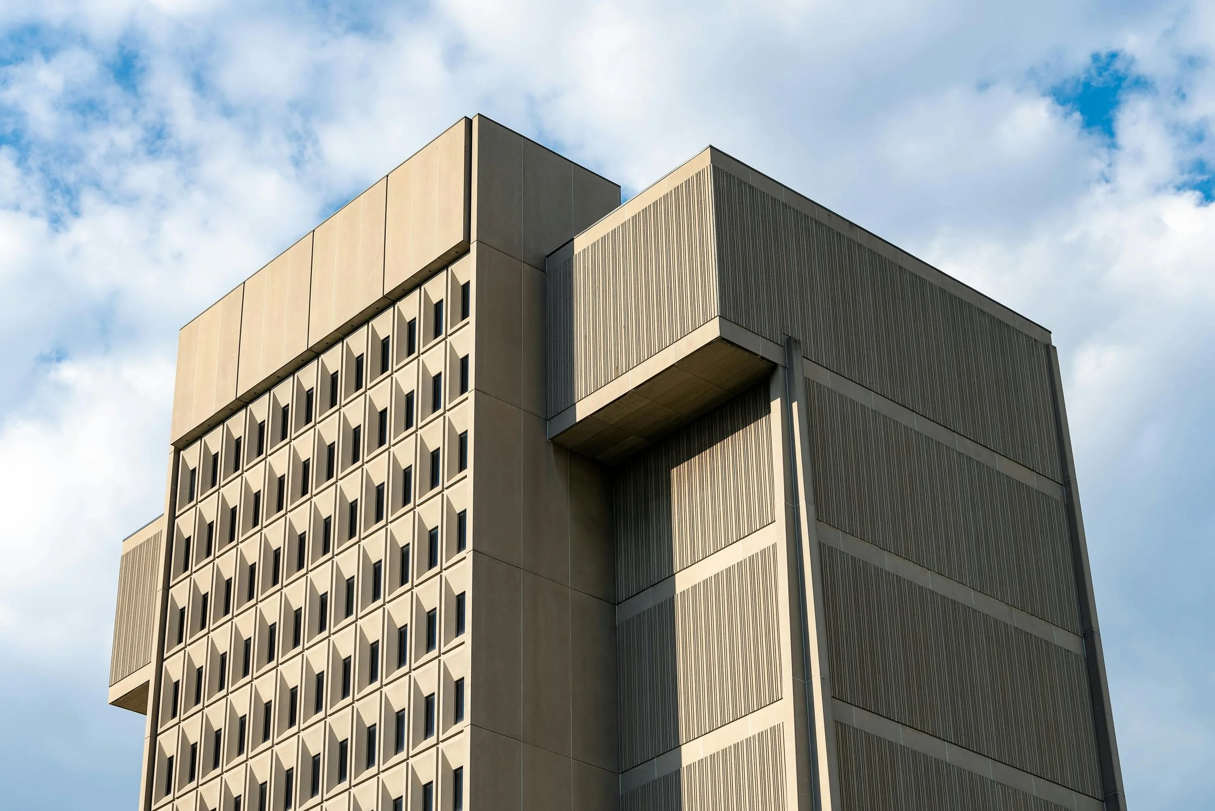 A modern multi-story building with a grid of small windows and concrete walls, under a partly cloudy sky.