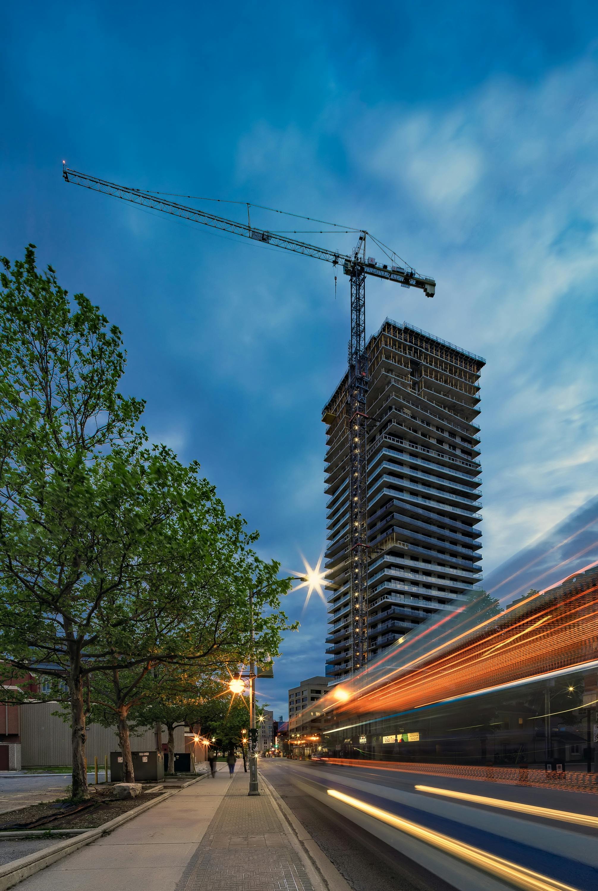 A city street scene at dusk featuring a high-rise building under construction with a crane on top, trees lining the sidewalk, streetlights, and blurred motion of a passing bus, with a partly cloudy sky in the background.