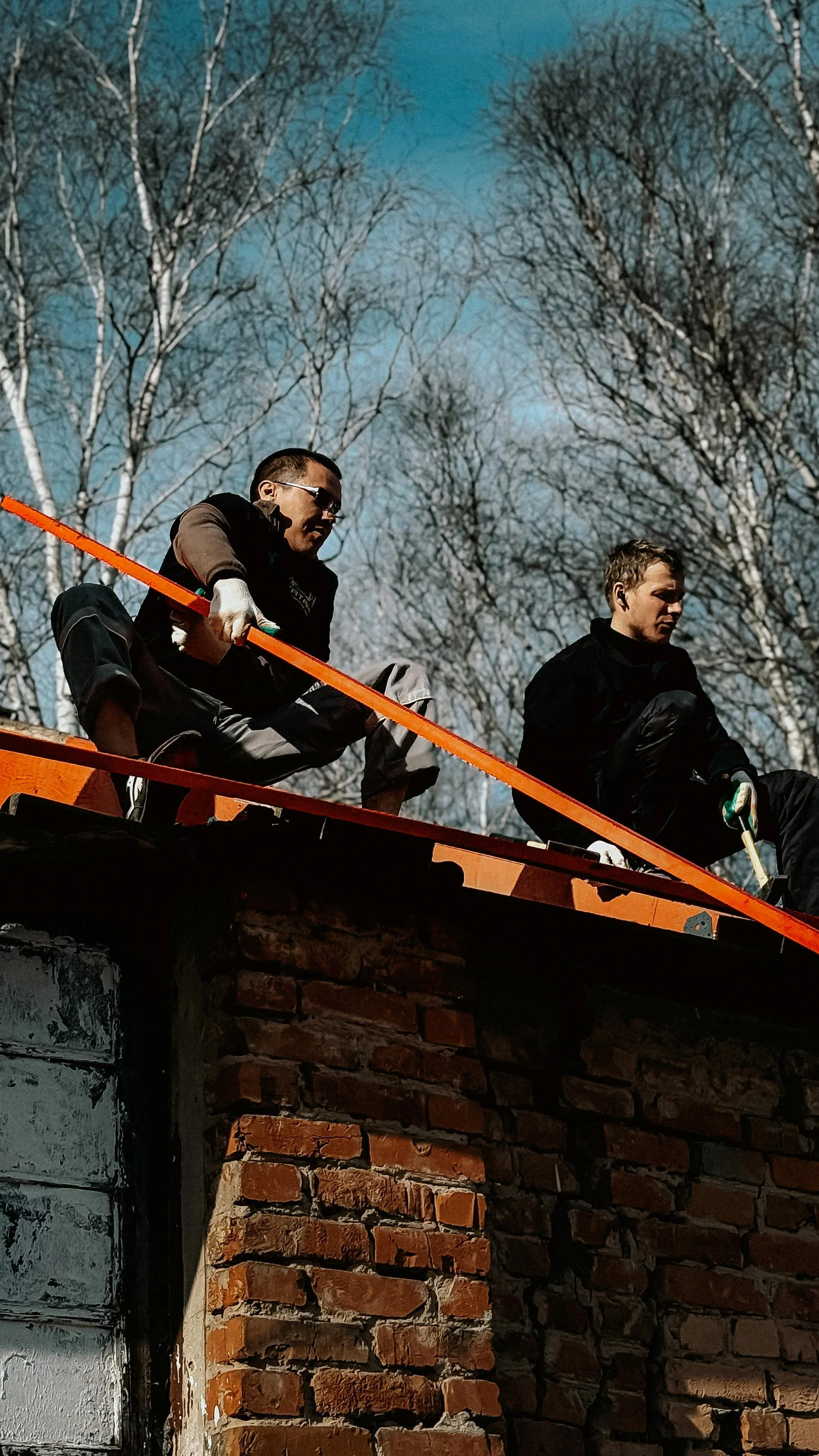 Two men working on a brick chimney on a rooftop, with leafless trees and a blue sky in the background.