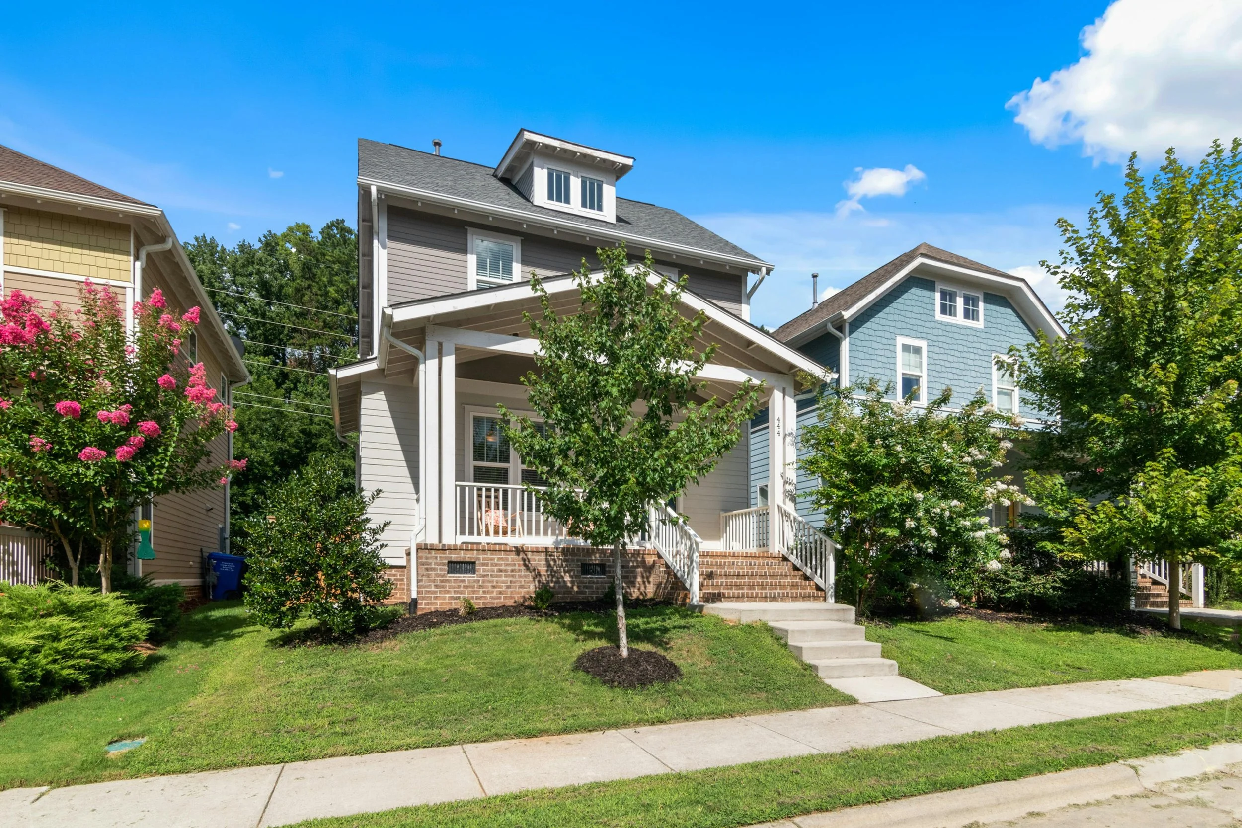 A residential neighborhood with three houses, front lawns, flowering trees, a sidewalk, and a clear blue sky with some clouds.