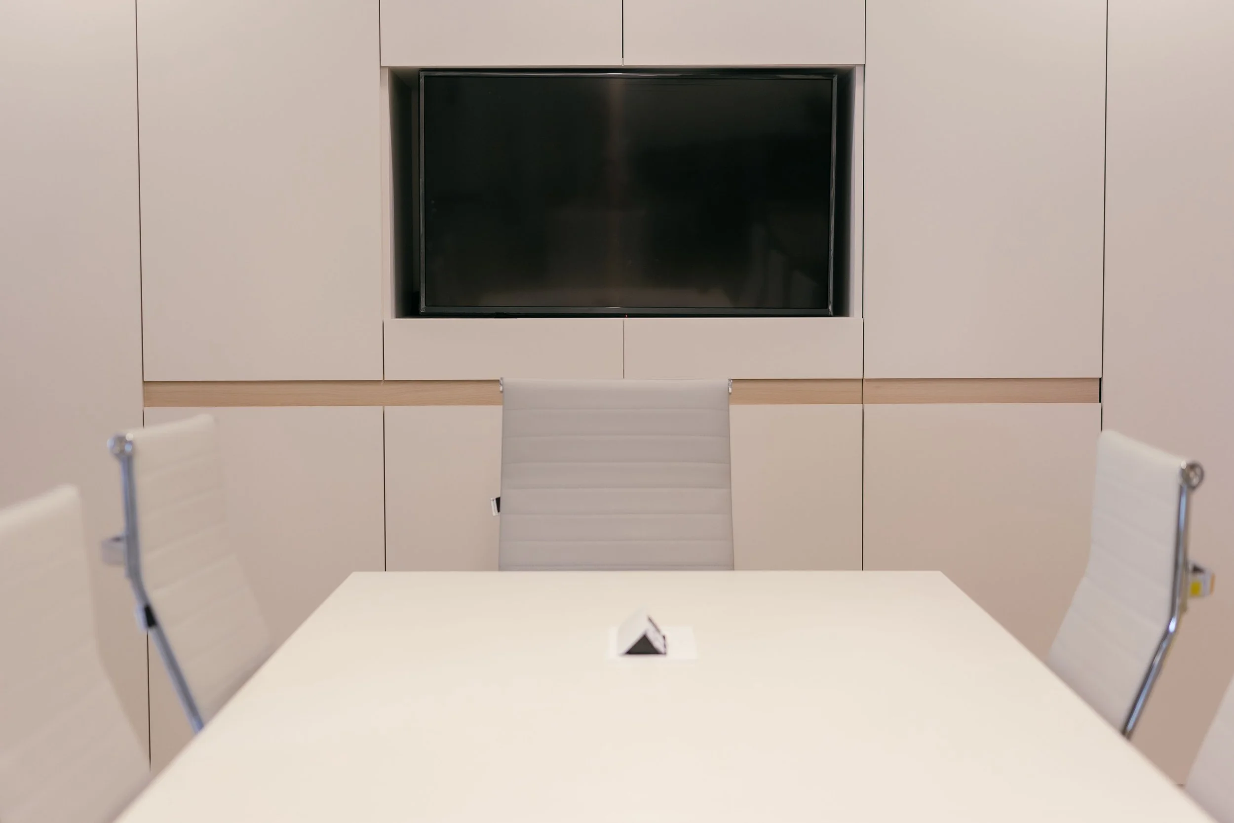 Modern conference room with a white table, white chairs, and a large flat-screen TV mounted on a white wall with built-in cabinets.