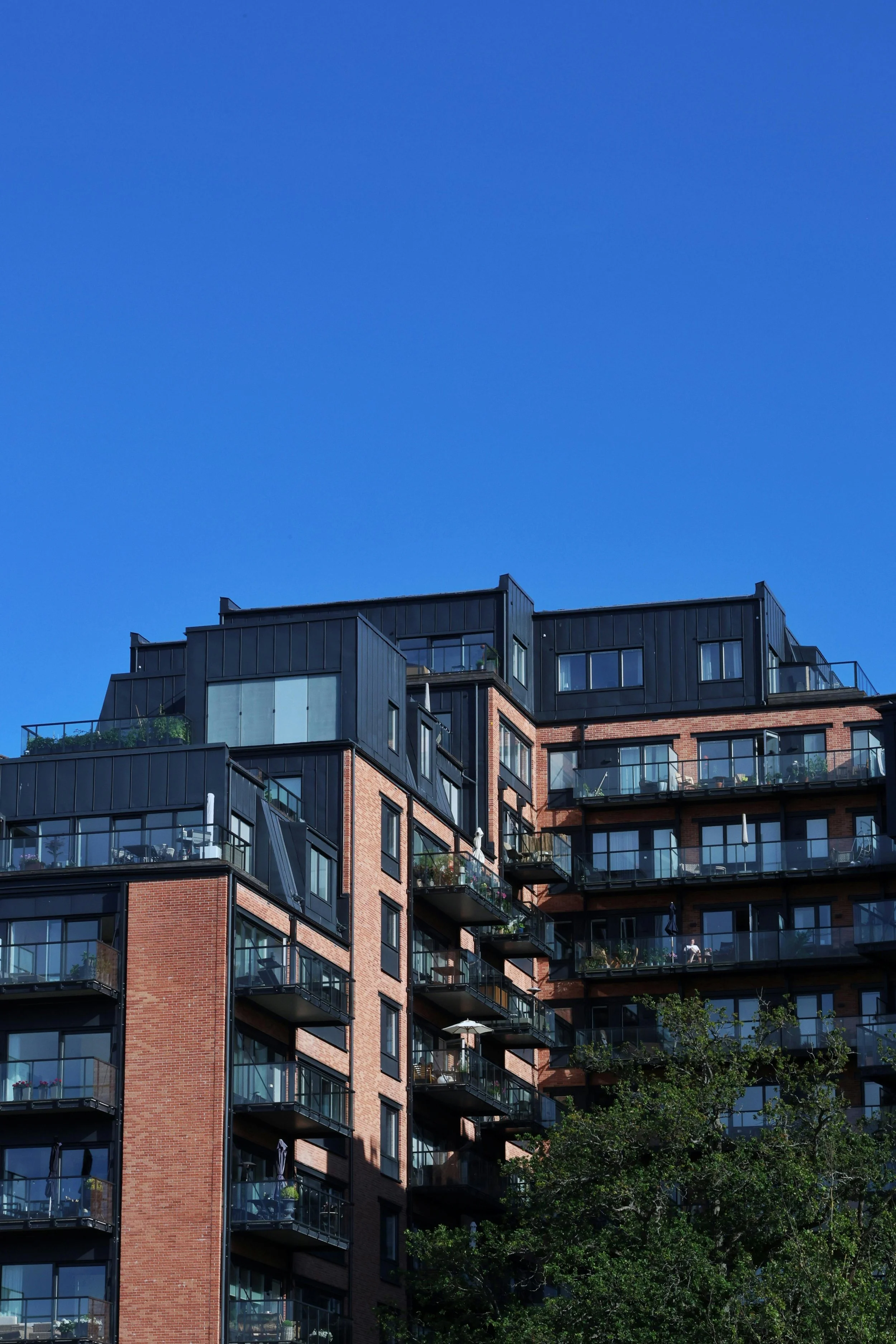 A tall residential building with brick and black metal exterior, multiple glass balconies, and large windows, set against a clear blue sky with some greenery at the bottom.