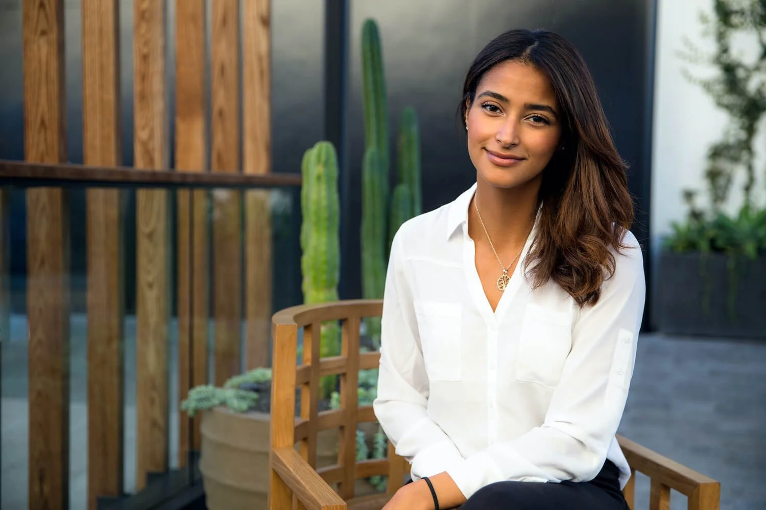 A woman with long dark hair sitting on a wooden chair outdoors, wearing a white blouse and a necklace, with a smile, in front of potted plants and a wooden fence.