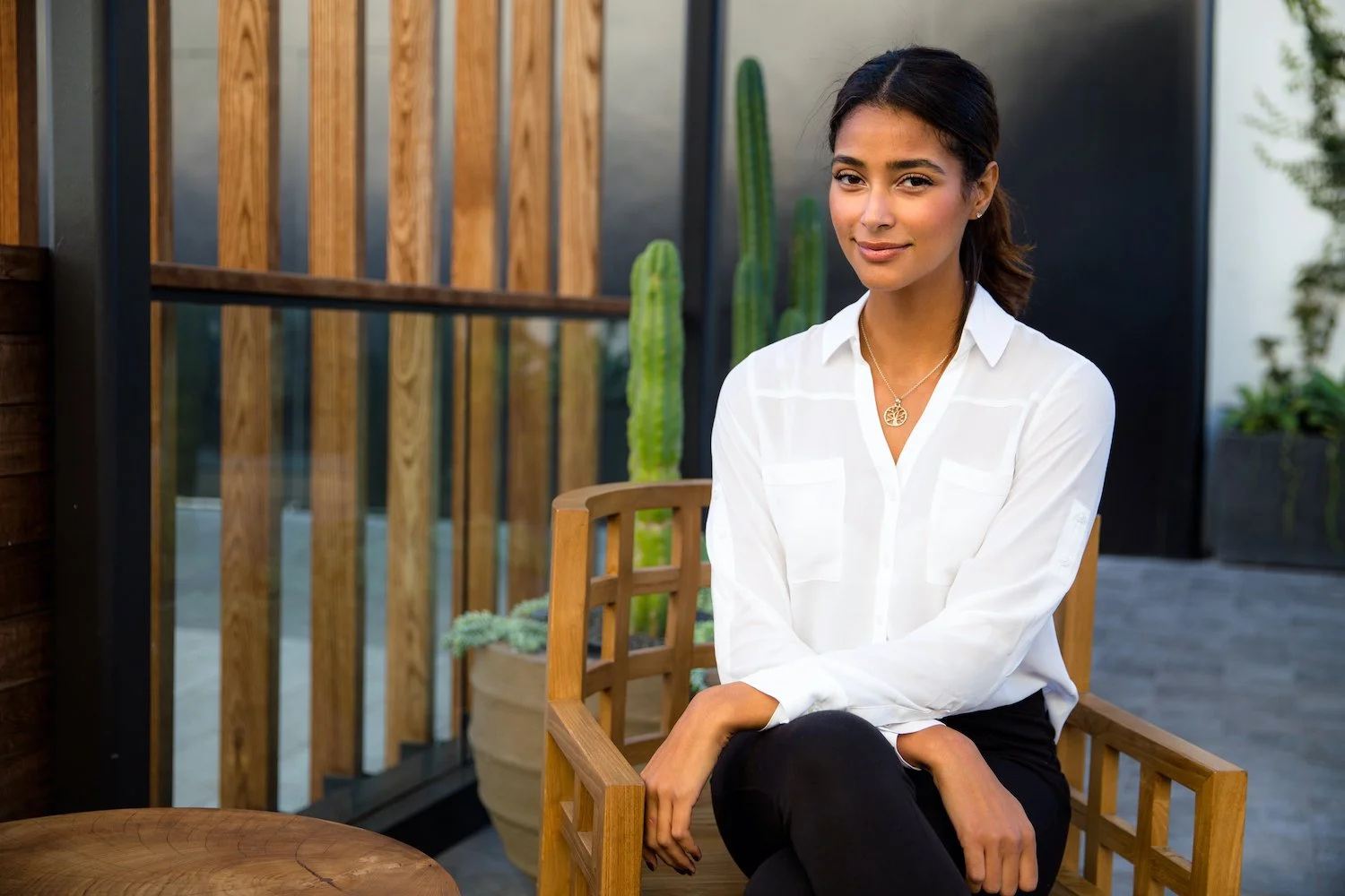 A young woman with dark hair, wearing a white shirt and black pants, sitting on a wooden chair outdoors with plants and wooden fencing in the background.