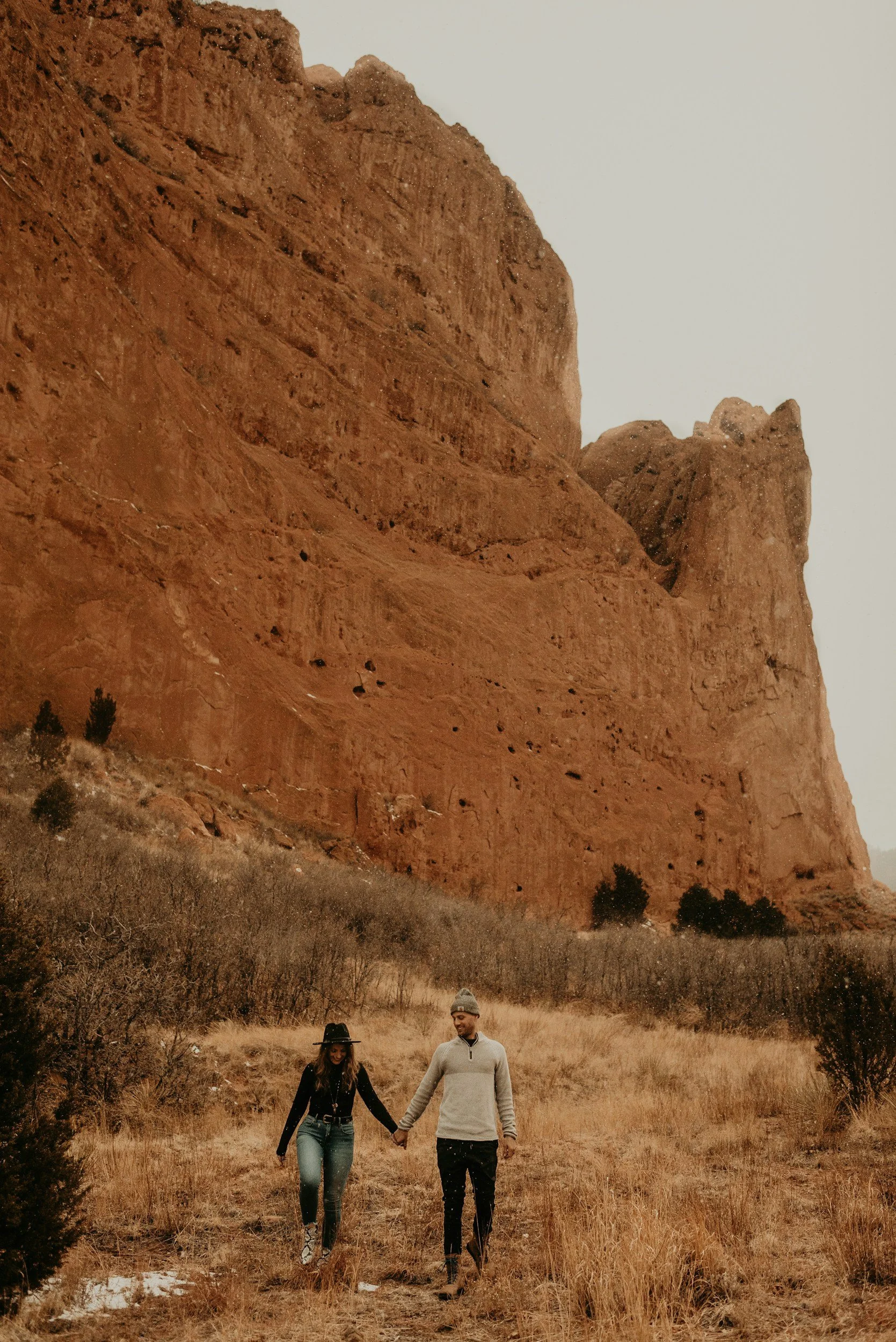 A couple holding hands and walking through a grassy field with large red rock formations in the background.