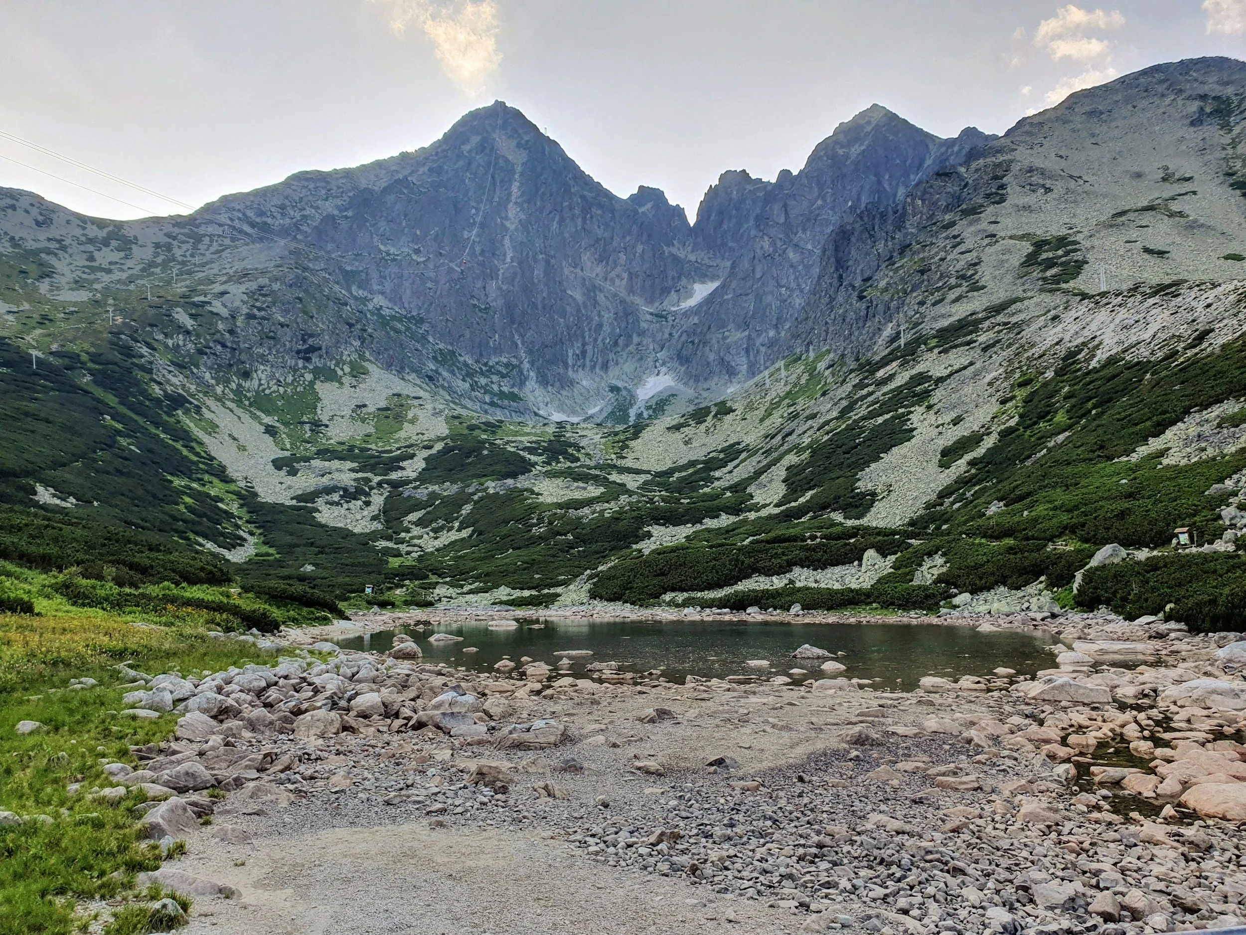 A mountain landscape with steep, rocky peaks, green slopes, a small lake, and a rocky shore in the foreground.