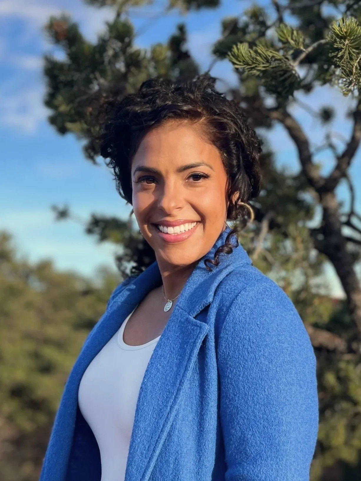A woman with dark curly hair smiling outdoors, wearing a blue jacket over a white top, with trees and a blue sky in the background.