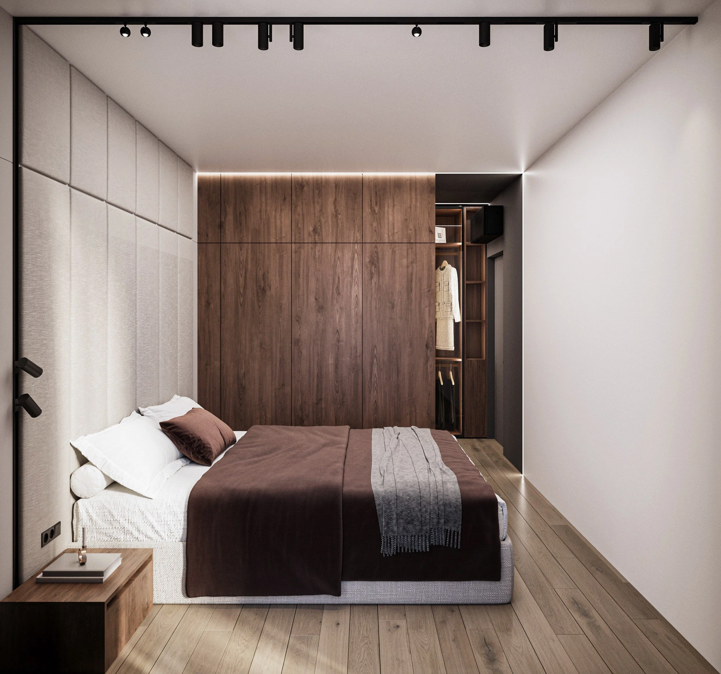 Modern bedroom with a bed featuring a brown bedspread, white pillows, and a gray throw. A wooden nightstand with books and a small clock is next to the bed. The room has a wooden wardrobe with sliding doors and a small open closet area with hanging c