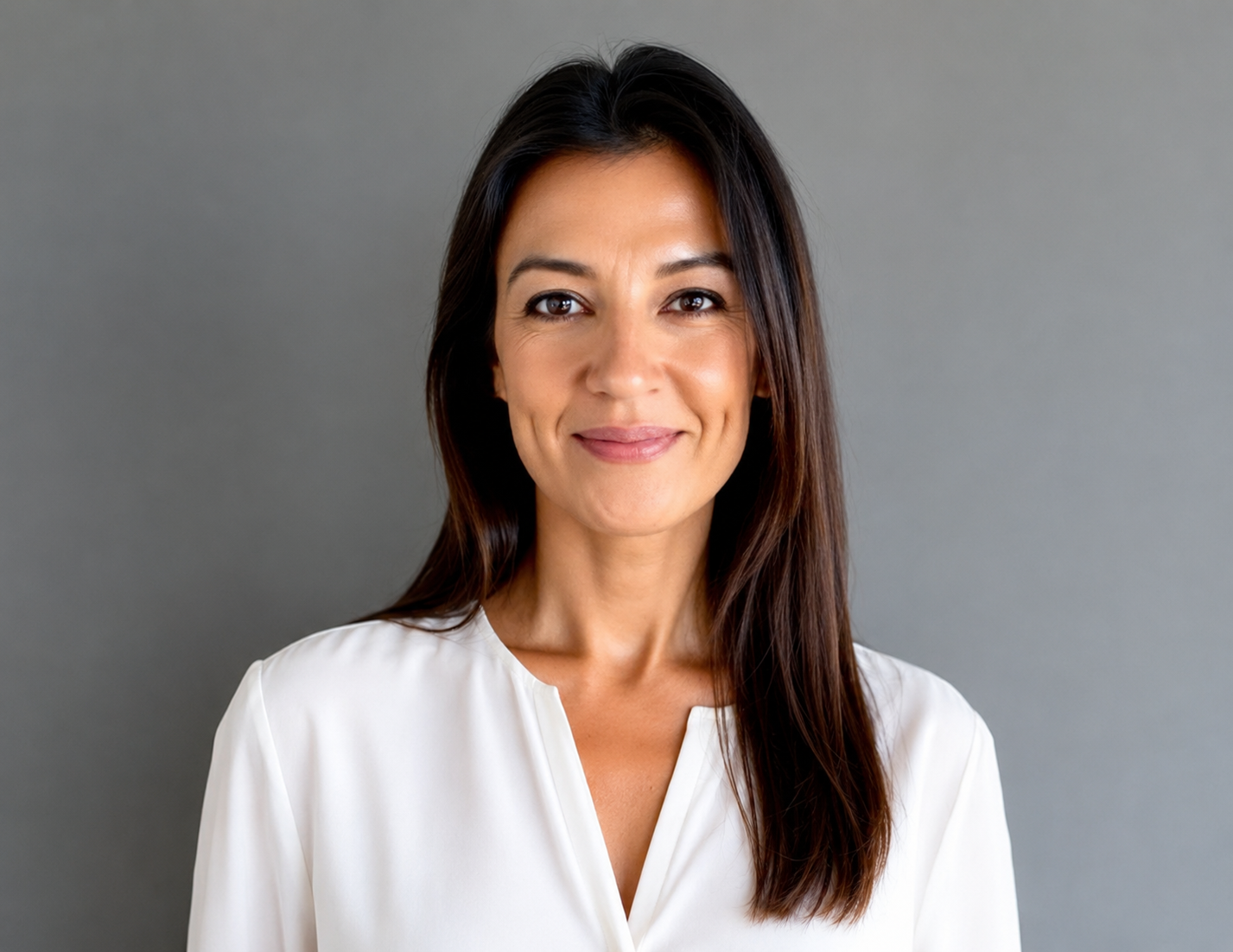 A woman with long dark hair wearing a white blouse, smiling at the camera, standing against a gray background.