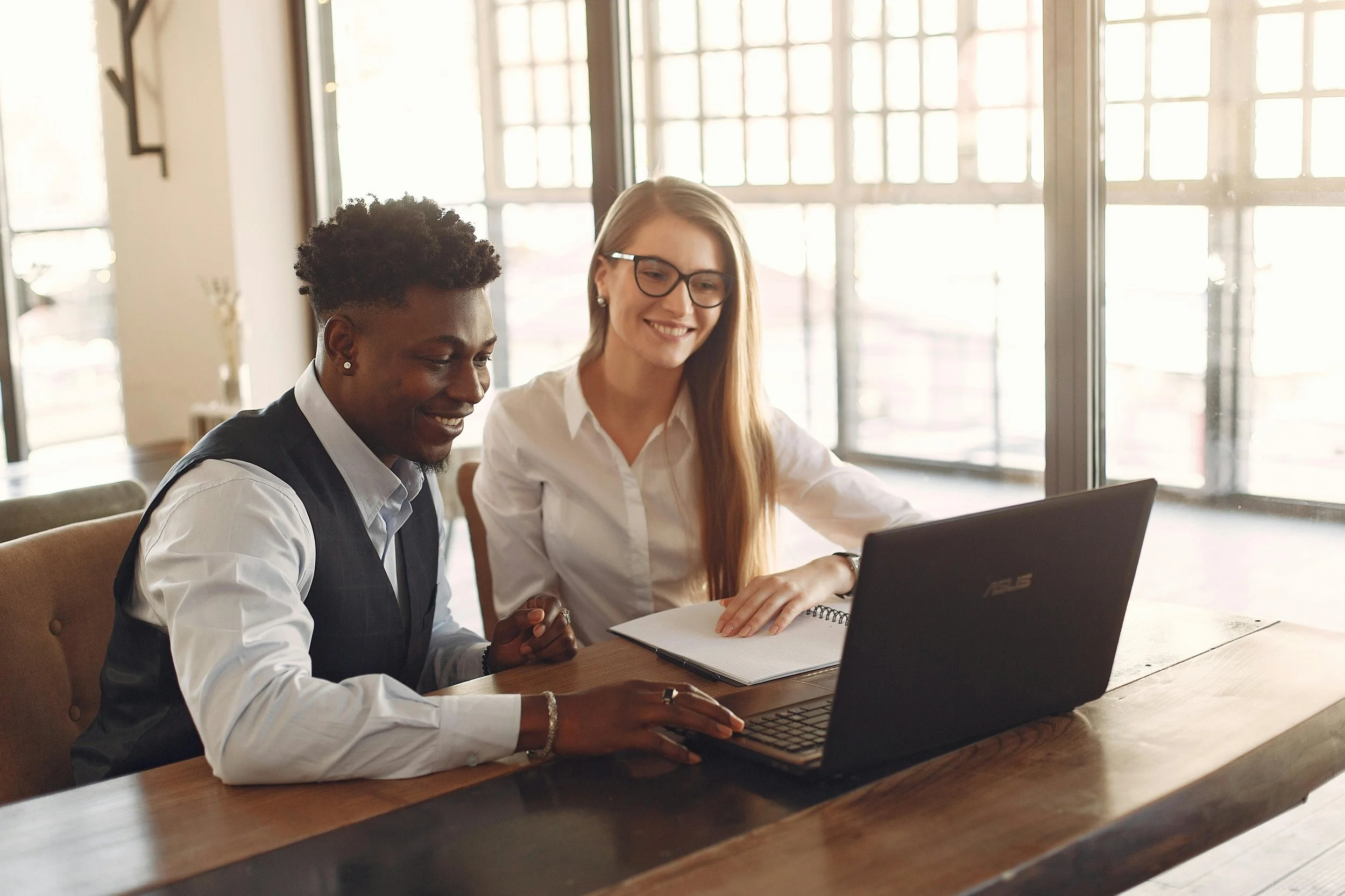 Two women sitting at a table, working on a laptop in a bright, modern office space.
