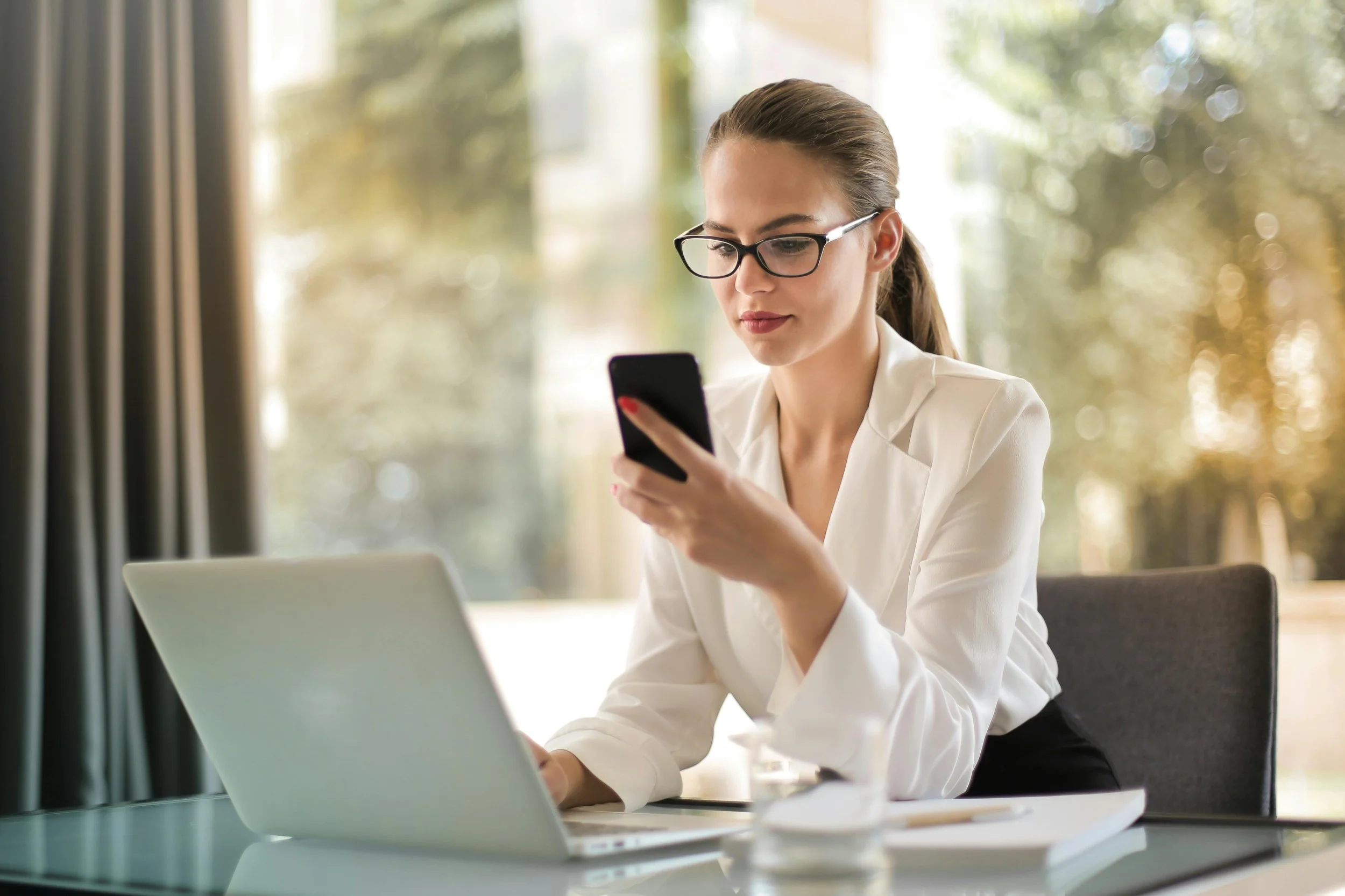 A woman with glasses and a white blouse is sitting at a desk, looking at her smartphone while working on a laptop.