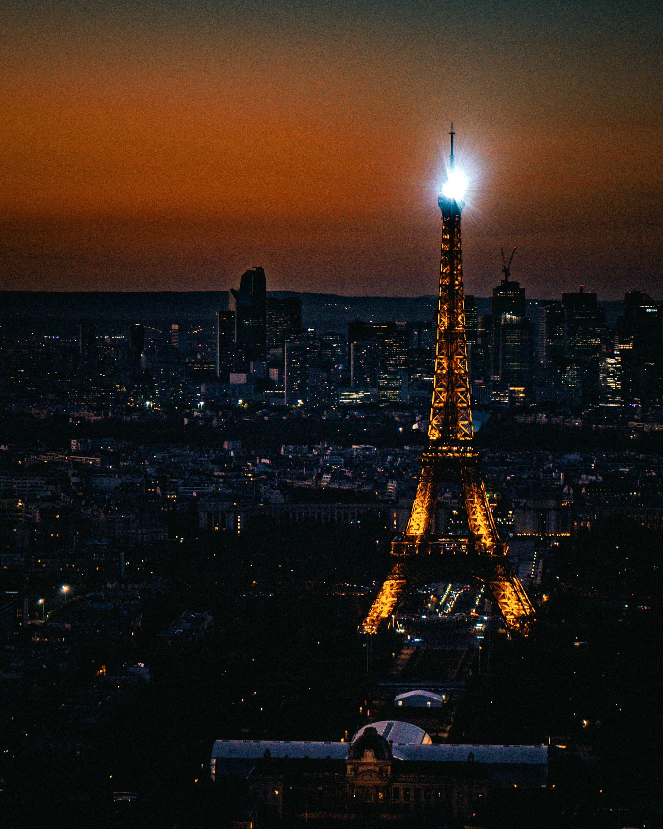 Tour Eiffel illuminée la nuit à Paris, avec un phare lumineux au sommet, surplombant la ville et le ciel orangeâtre.