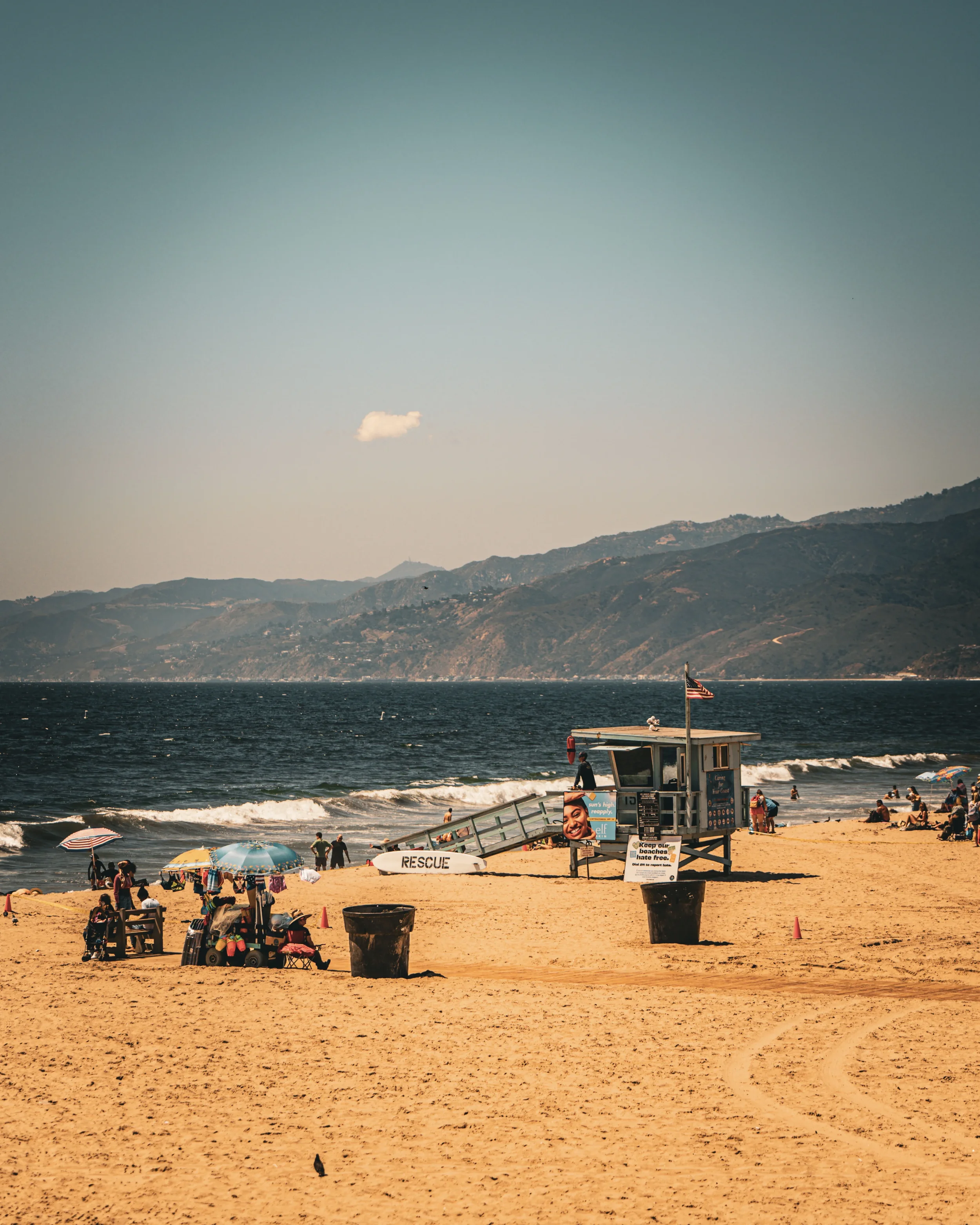 Plage de sable avec des parasols, un poste de sauveteurs, des personnes au bord de la mer, des montagnes en arrière-plan et un ciel clair.