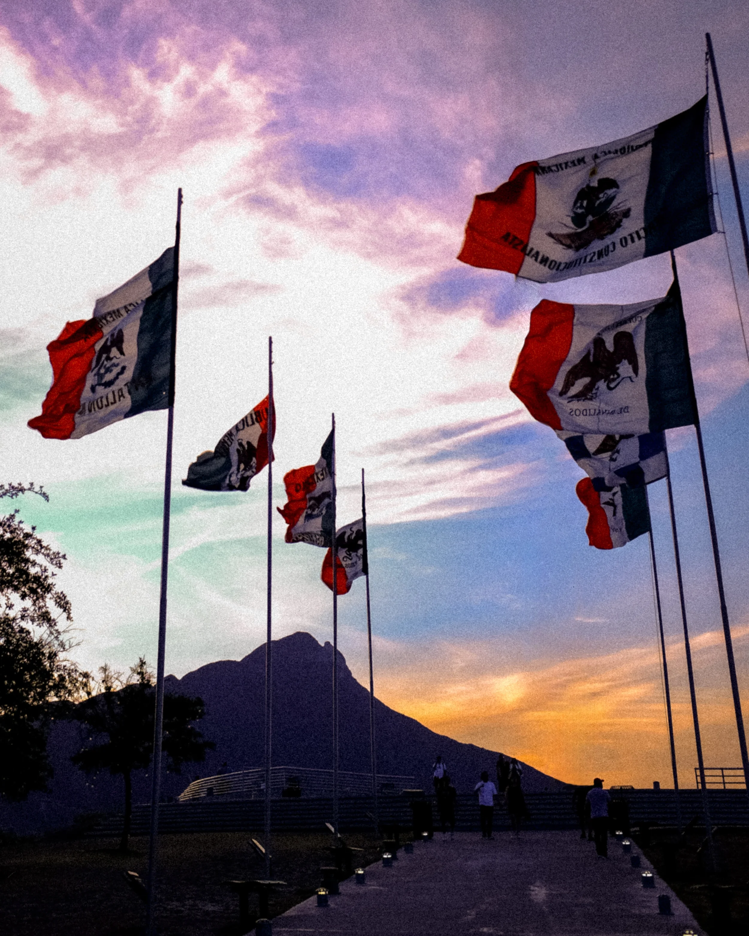 Drapeaux français flottant au vent lors d'un coucher de soleil, avec une montagne en arrière-plan et des personnes marchant sur un chemin dans un parc.