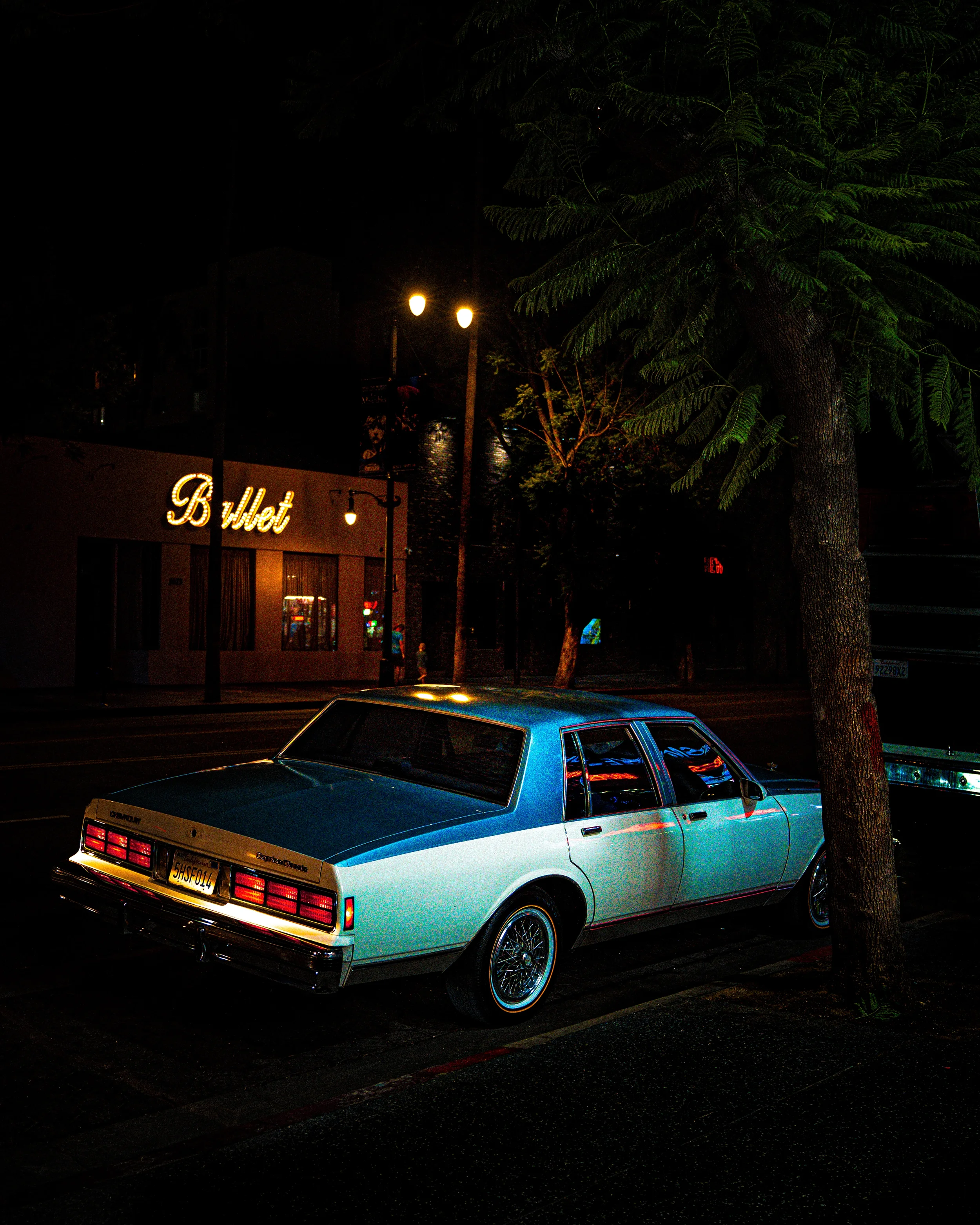 Une voiture ancienne bleue garée près d'un arbre, de nuit, avec un éclairage urbain et un panneau lumineux 'Ballet' en arrière-plan.