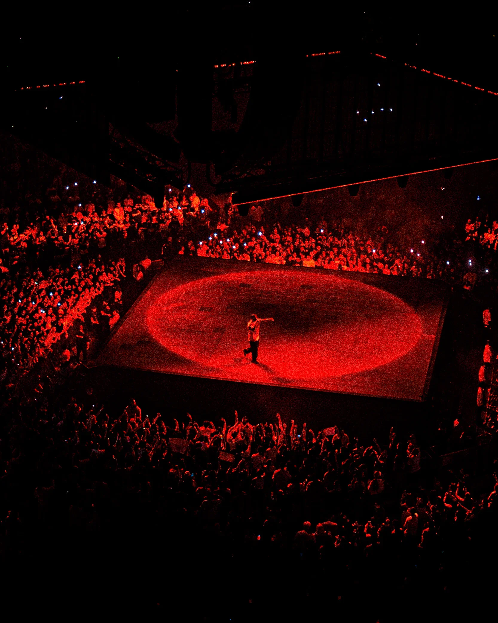 Un artiste chante sur une scène de cirque sous un éclairage rouge, entouré d'un public nombreux dans une arène.